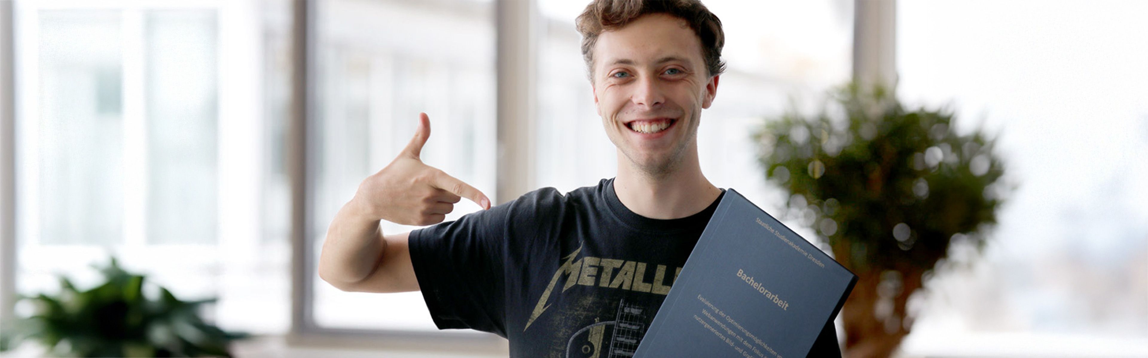 Eine lächelnde Person in einem schwarzen T-Shirt zeigt auf ein Dokument mit blauem Umschlag und steht in einem hellen Raum mit Pflanzen im Hintergrund.