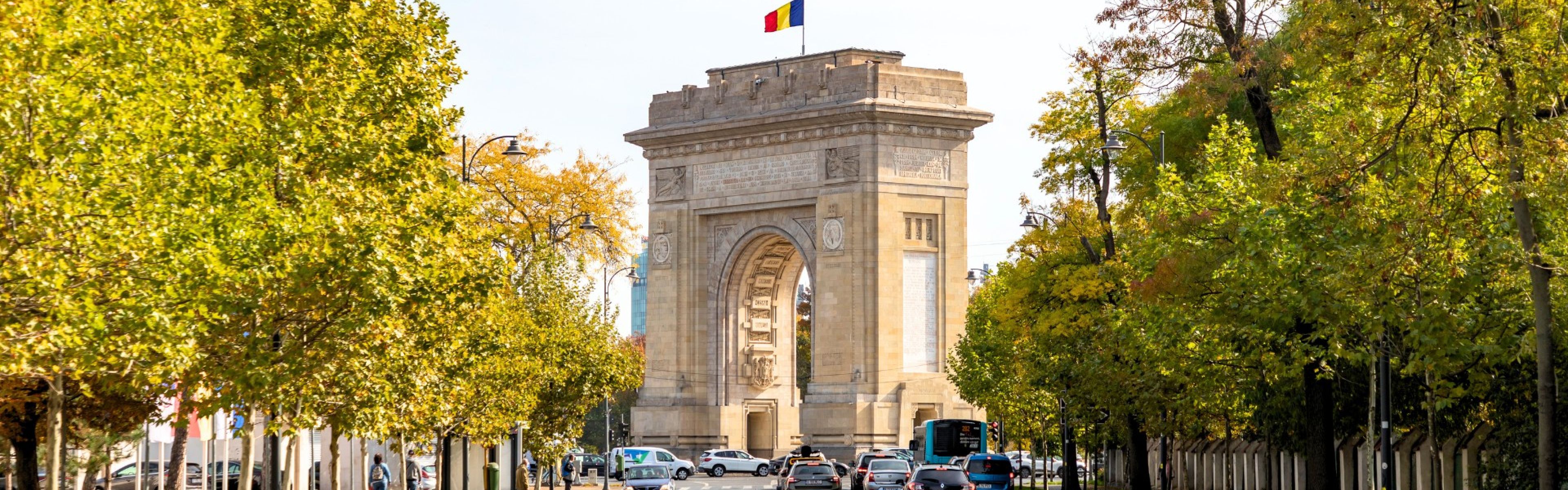 Triumphbogen mit einer Fahne auf der Spitze, umgeben von Bäumen und einer belebten Straße mit Autos, unter einem klaren Himmel.