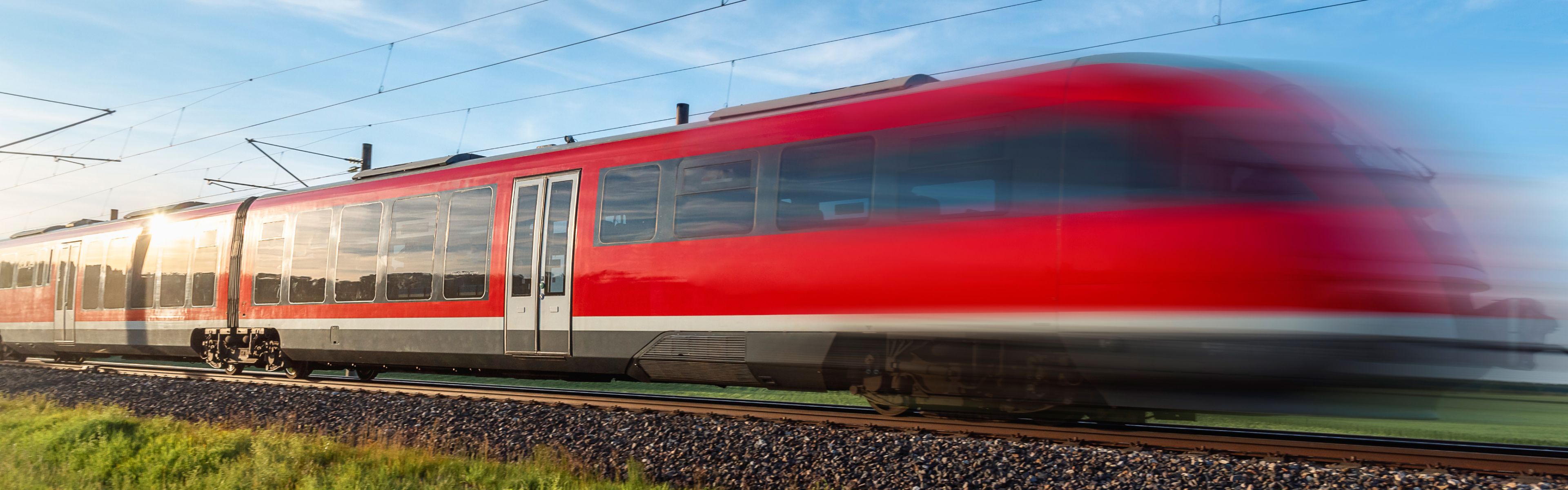 ChatGPT: Ein roter Zug fährt mit hoher Geschwindigkeit über eine Bahnstrecke in ländlicher Umgebung, wobei die Bewegungsunschärfe auf das hohe Tempo hinweist. Der Himmel ist klar und blau.