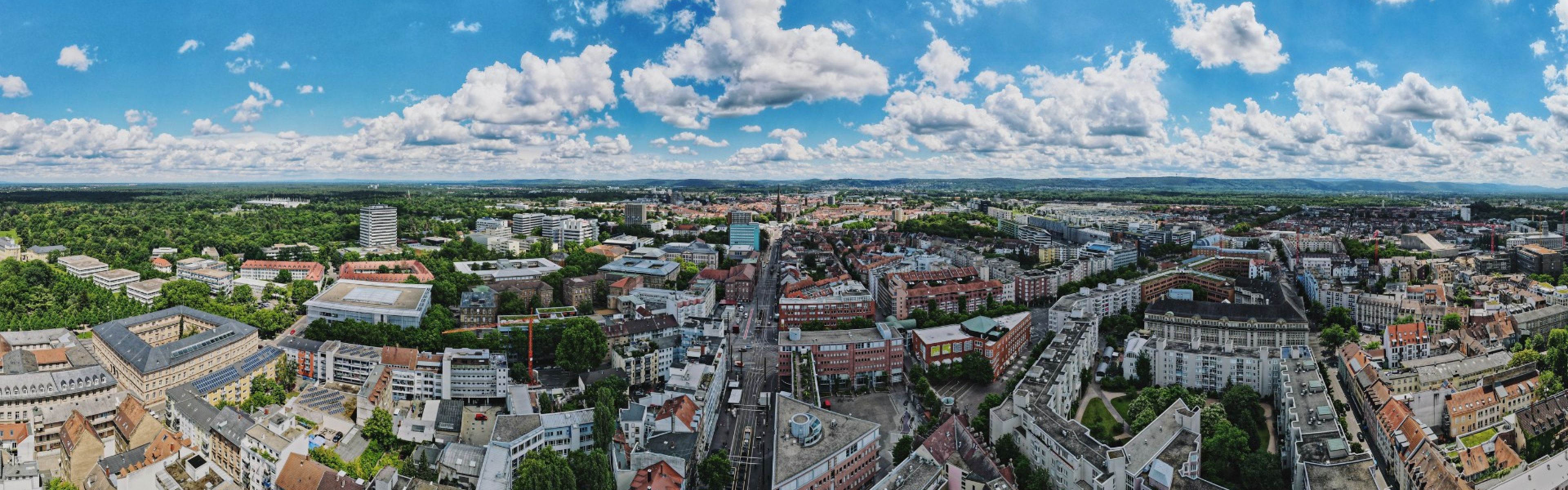 Panoramablick auf eine Stadtlandschaft mit einer Mischung aus modernen und traditionellen Gebäuden, umgeben von üppigem Grün unter einem teilweise bewölkten blauen Himmel.