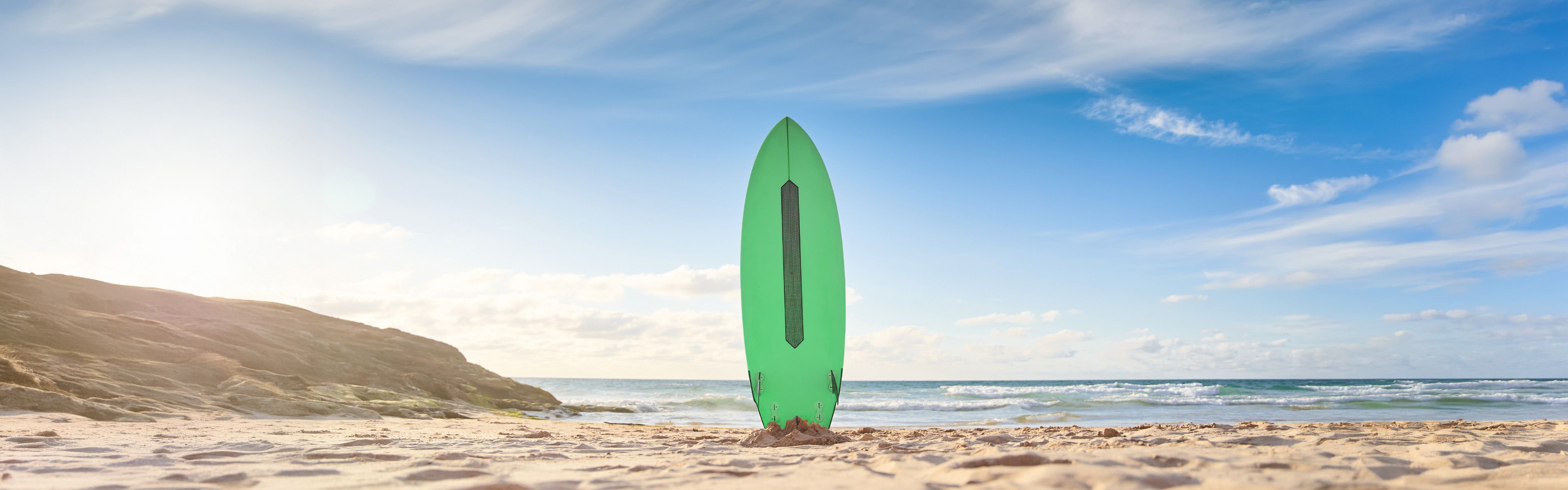 Ein grünes Surfbrett steht aufrecht an einem Sandstrand, im Hintergrund das Meer und ein klarer blauer Himmel.