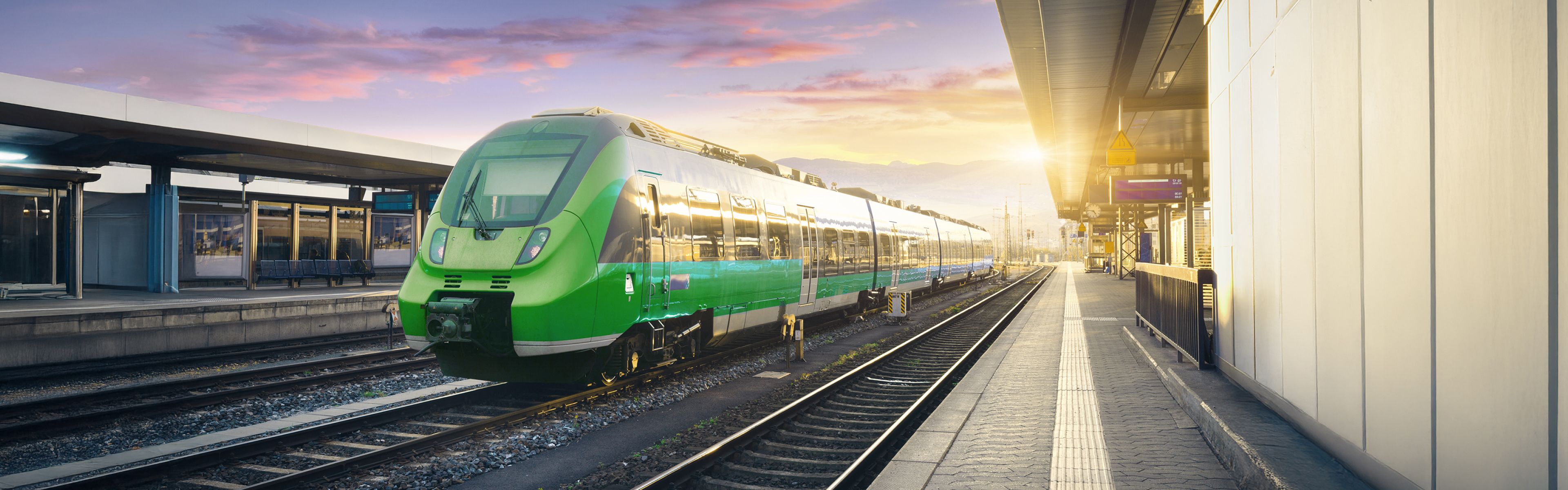 Grüner Zug an einem modernen Bahnsteig während des Sonnenuntergangs, mit klarem Himmel und leuchtendem Sonnenlicht im Hintergrund.