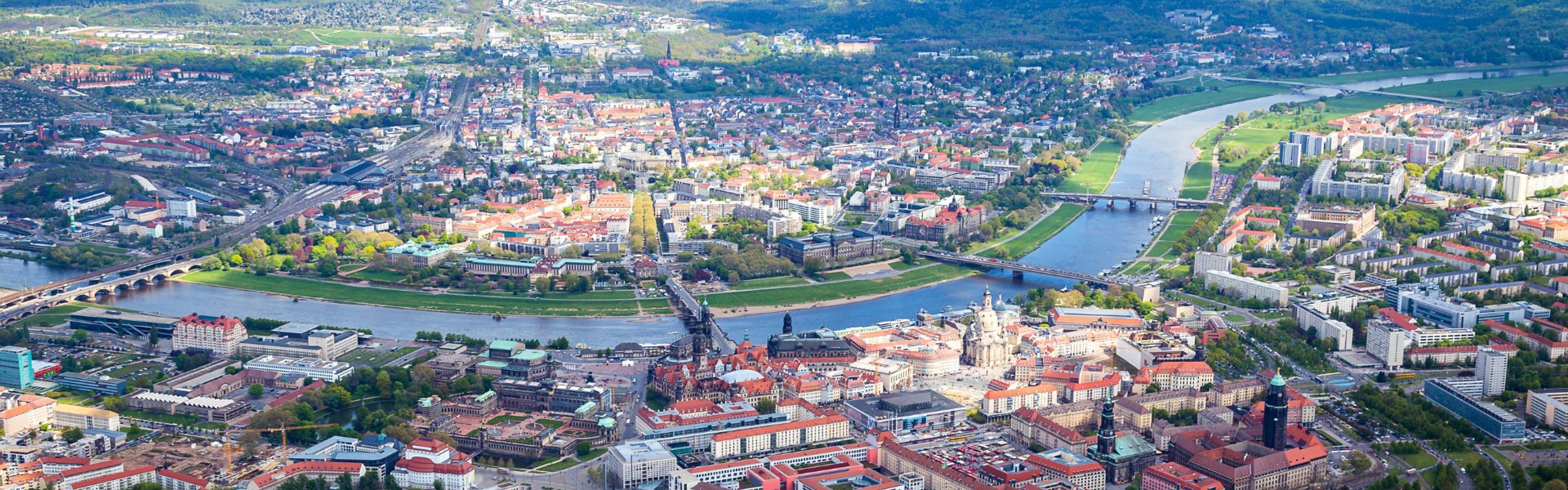 Draufsicht auf eine Stadt mit einem gewundenen Fluss, Brücken und einer Mischung aus historischen und modernen Gebäuden, umgeben von üppigem Grün und fernen Hügeln.