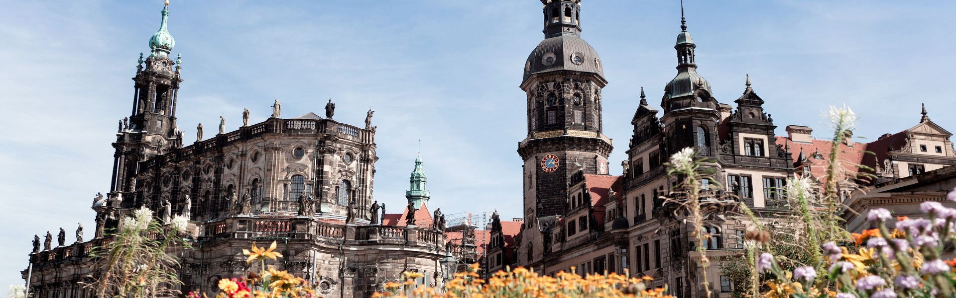 Historische europäische Gebäude mit verzierten Türmen und Spitzen vor blauem Himmel, mit bunten Blumen im Vordergrund.