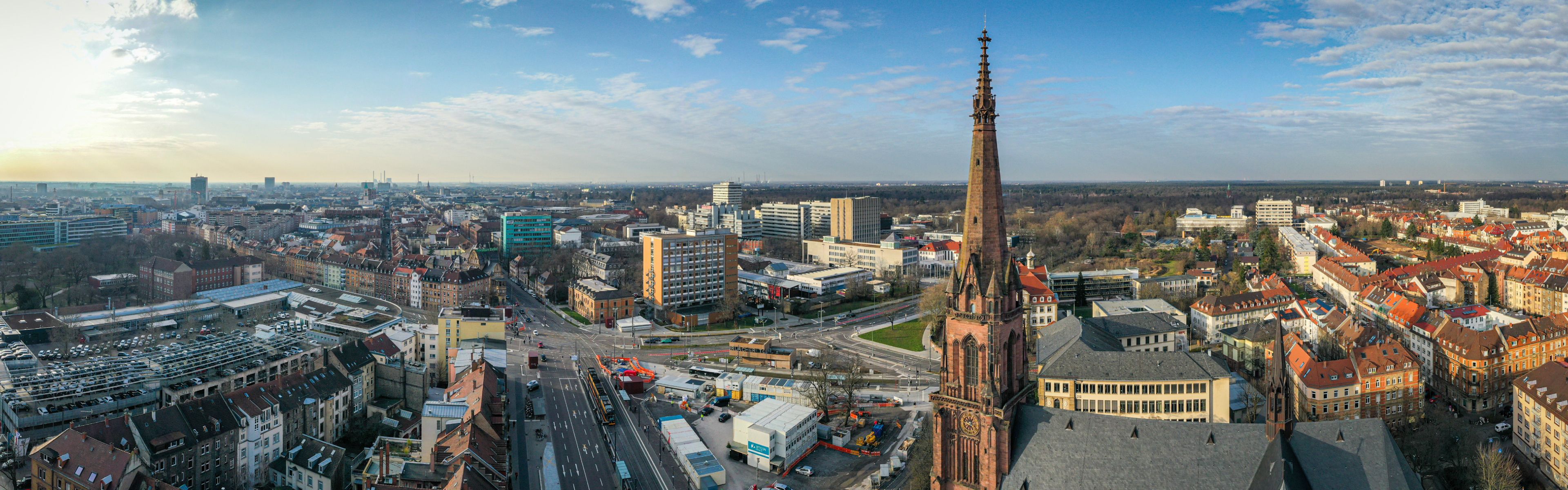 Panoramablick auf eine Stadtansicht mit einem markanten Kirchturm, umgeben von Gebäuden, unter einem klaren blauen Himmel.