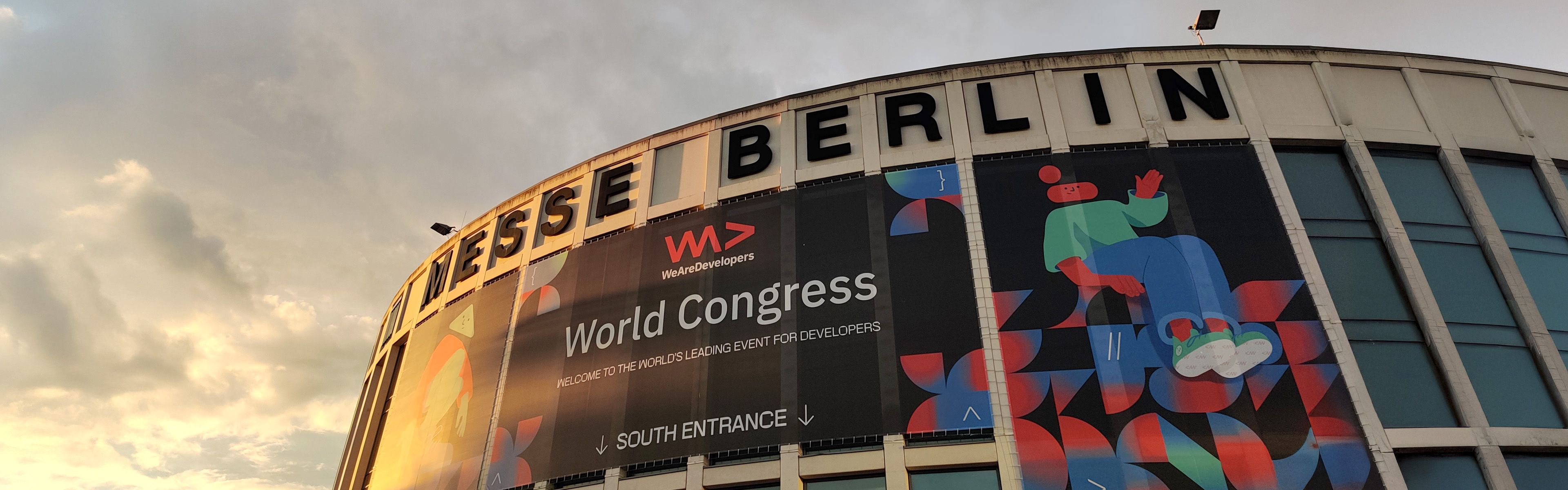 Außenansicht der Messe Berlin mit einem großen Banner für den Weltkongress, das farbenfrohe abstrakte Designs zeigt, vor einem Himmel im Sonnenuntergang.