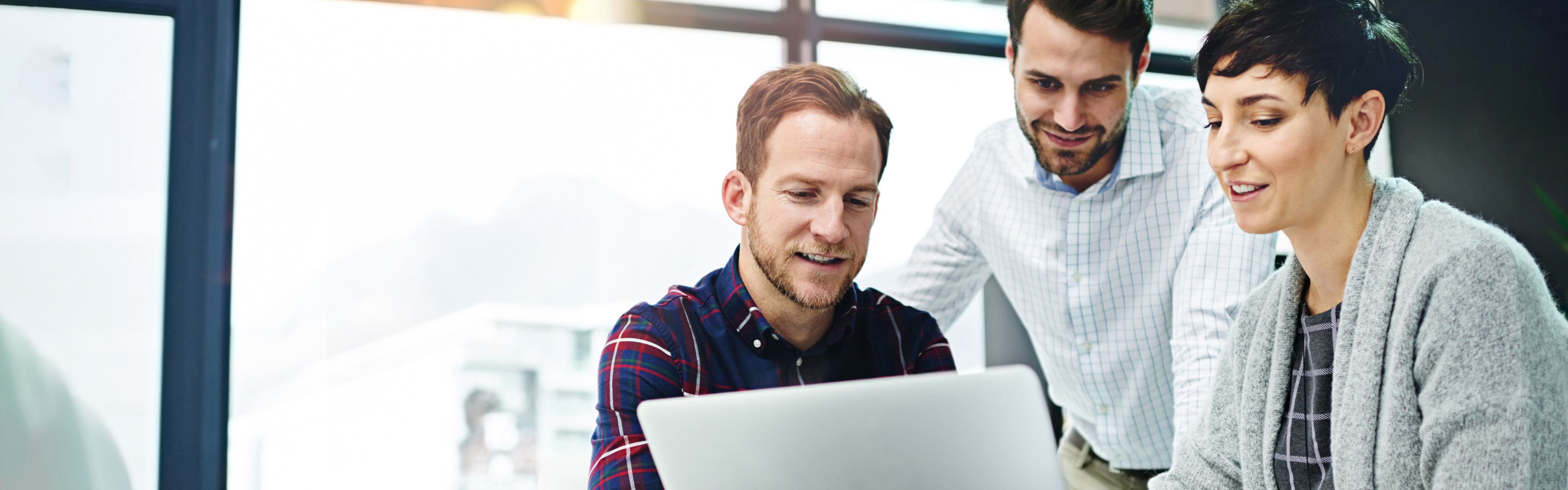 Drei Kollegen arbeiten gemeinsam an einem Laptop in einem modernen Büro. Ein Mann im karierten Hemd sitzt am Laptop, während die anderen beiden daneben stehen und auf den Bildschirm schauen