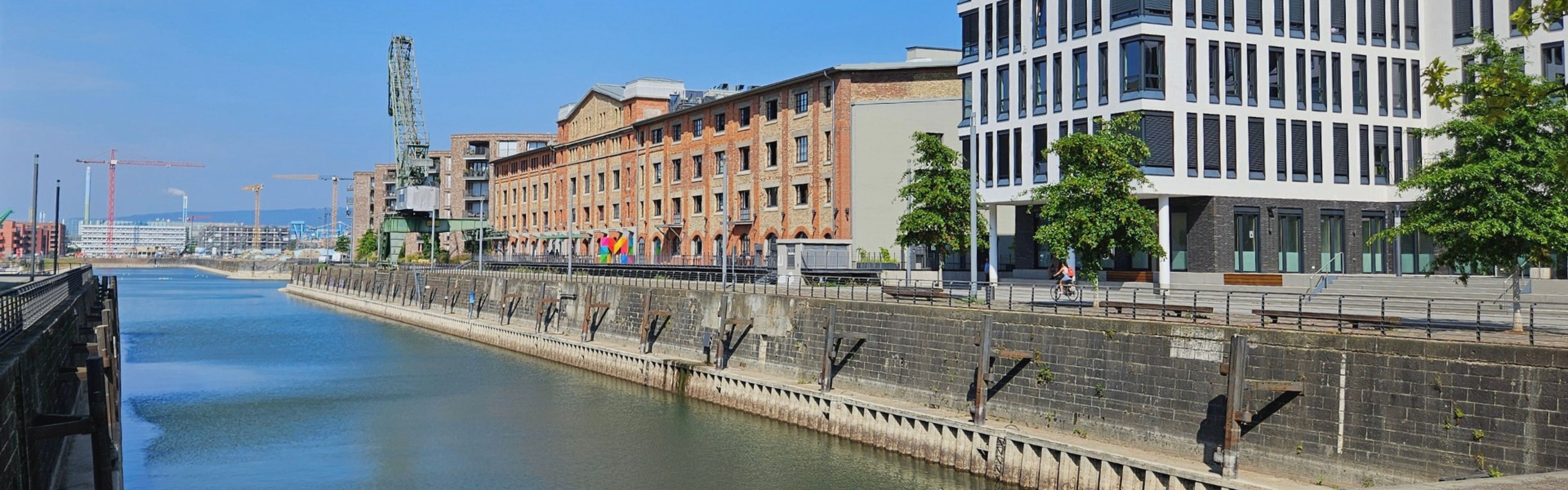 Kanal, gesäumt von modernen und historischen Gebäuden, Bäumen und einem klaren blauen Himmel.