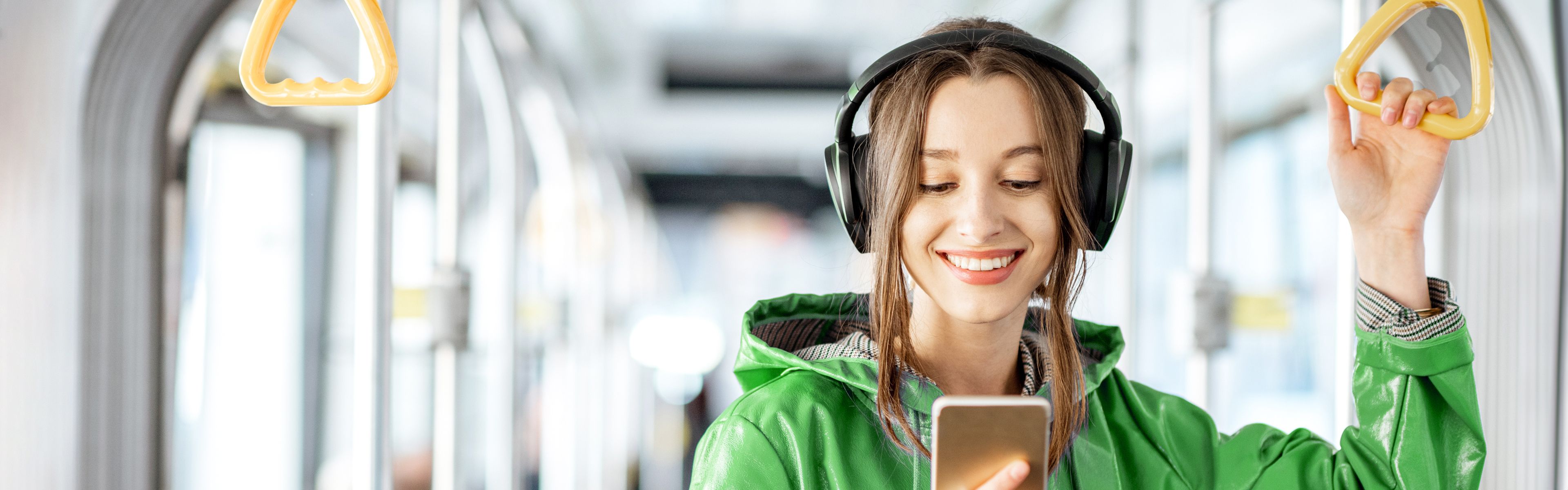 Eine lächelnde Frau in einer grünen Jacke hört Musik mit Kopfhörern und hält ein Smartphone in der Hand, während sie in einem Bus eine gelbe Haltestange festhält.