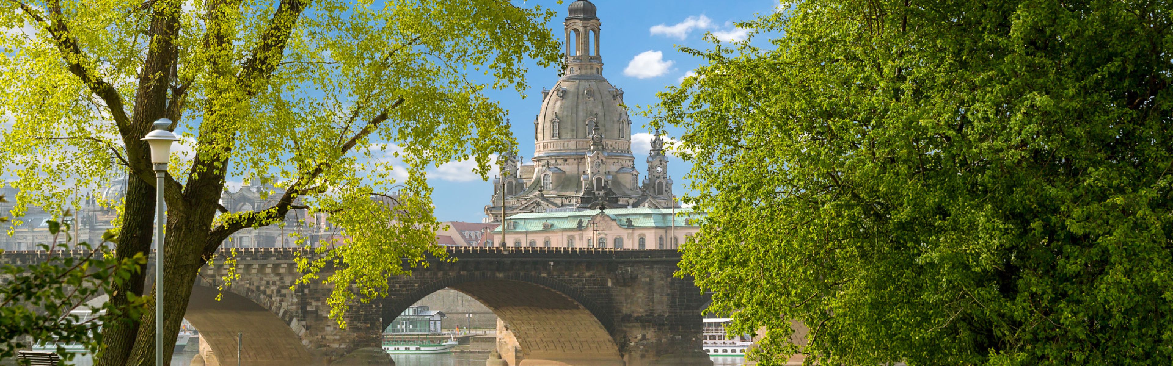 Historische Brücke mit kunstvoller Architektur, eingerahmt von üppig grünen Bäumen unter einem blauen Himmel mit vereinzelten Wolken.