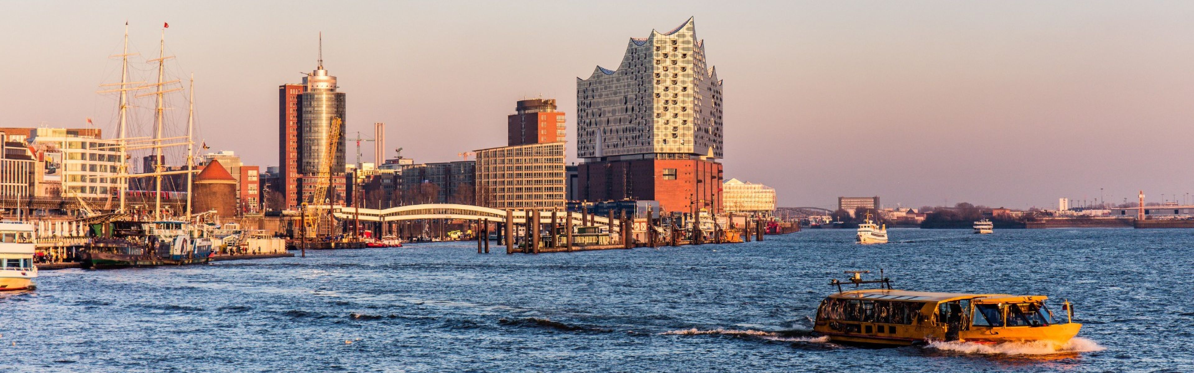 Stadtbild am Wasser mit modernen und historischen Gebäuden, darunter eine markante Glasstruktur. Boote befahren den Fluss unter einem klaren Himmel.