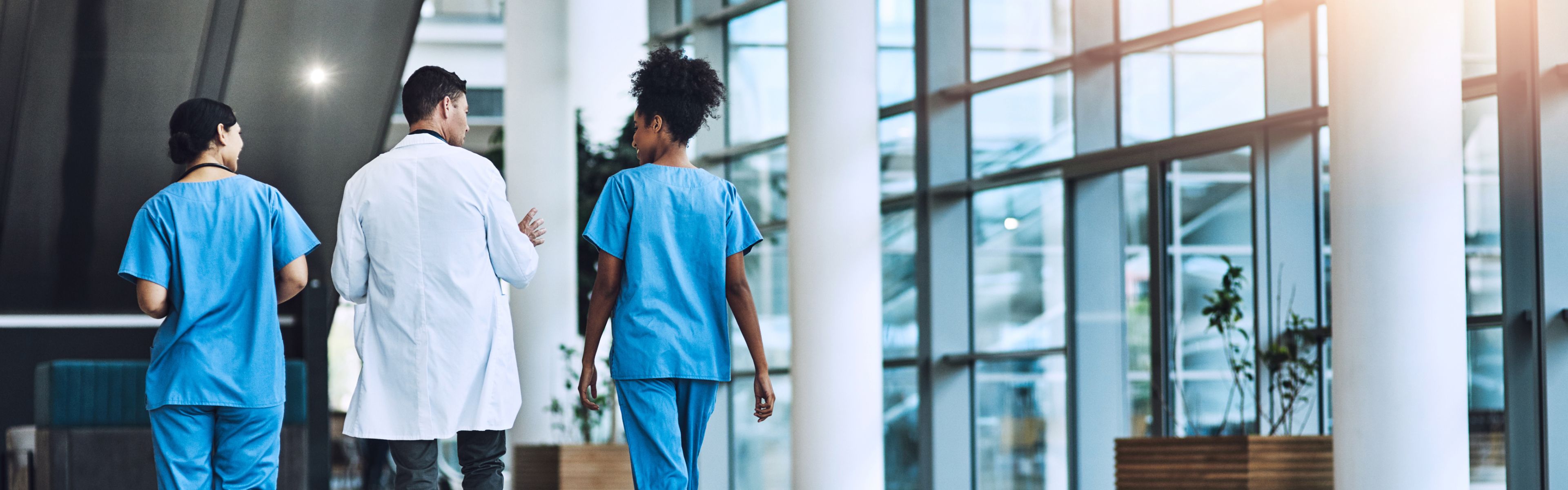 Drei medizinische Fachkräfte gehen einen Flur mit großen Fenstern entlang, zwei in blauer Arbeitskleidung und eine in weißem Kittel, vertieft in ein Gespräch.