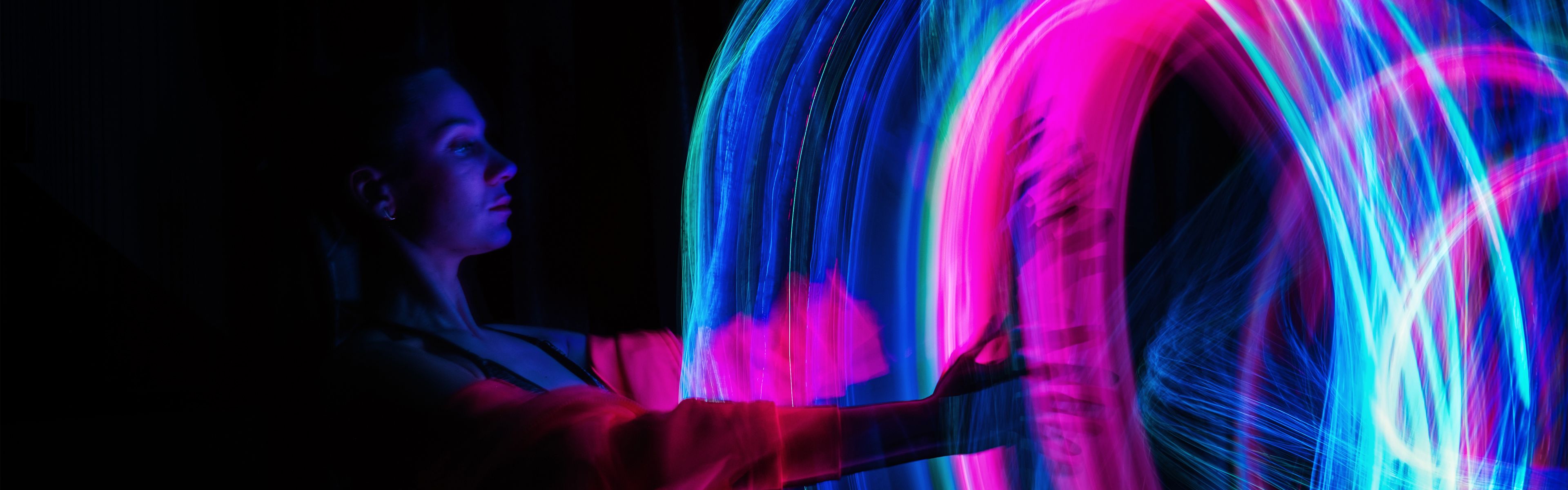 Eine Person interagiert mit lebendigen, wirbelnden Neonlichtern in einem dunklen Raum, wodurch ein dynamischer und farbenfroher visueller Effekt entsteht.