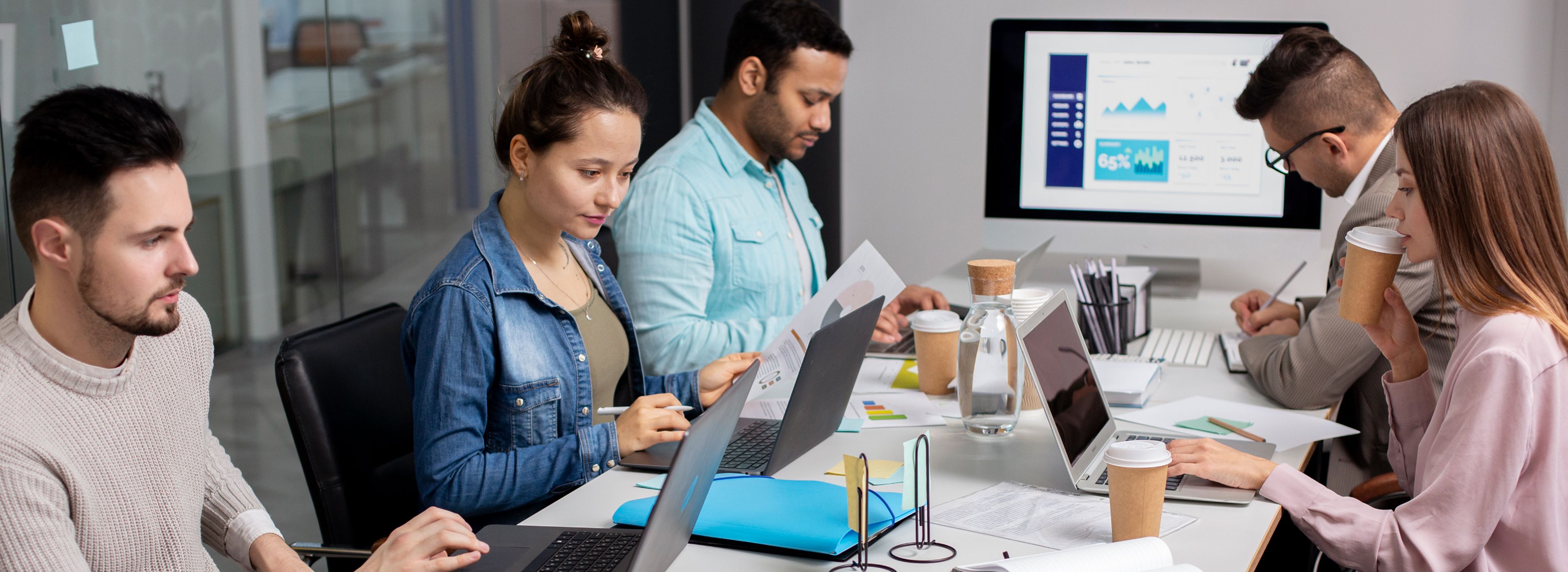 Fünf Personen arbeiten an einem Tisch mit Laptops, Papieren und Kaffeetassen. Im Hintergrund zeigt ein Bildschirm Grafiken und Diagramme.