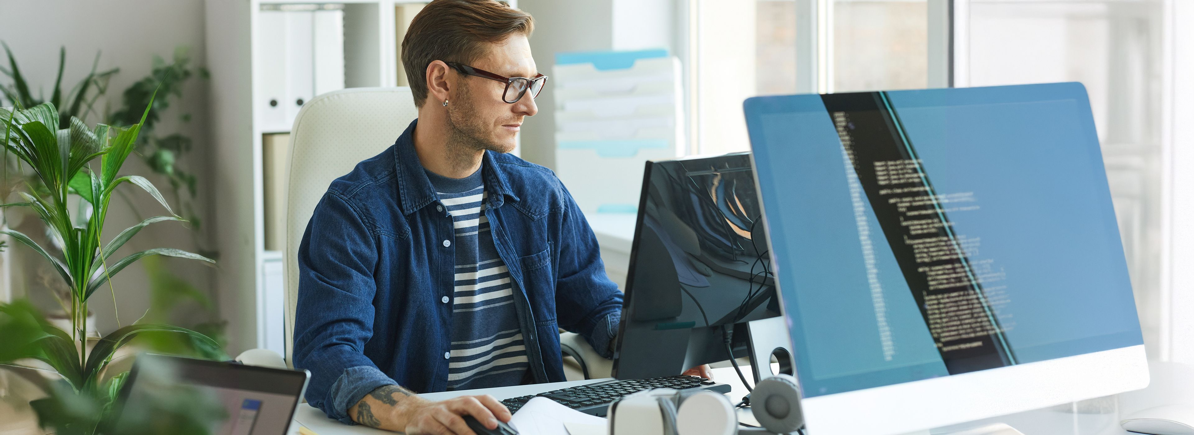 Eine Person arbeitet an einem Schreibtisch mit zwei Computerbildschirmen, auf denen Code angezeigt wird, in einer hellen Büroumgebung.