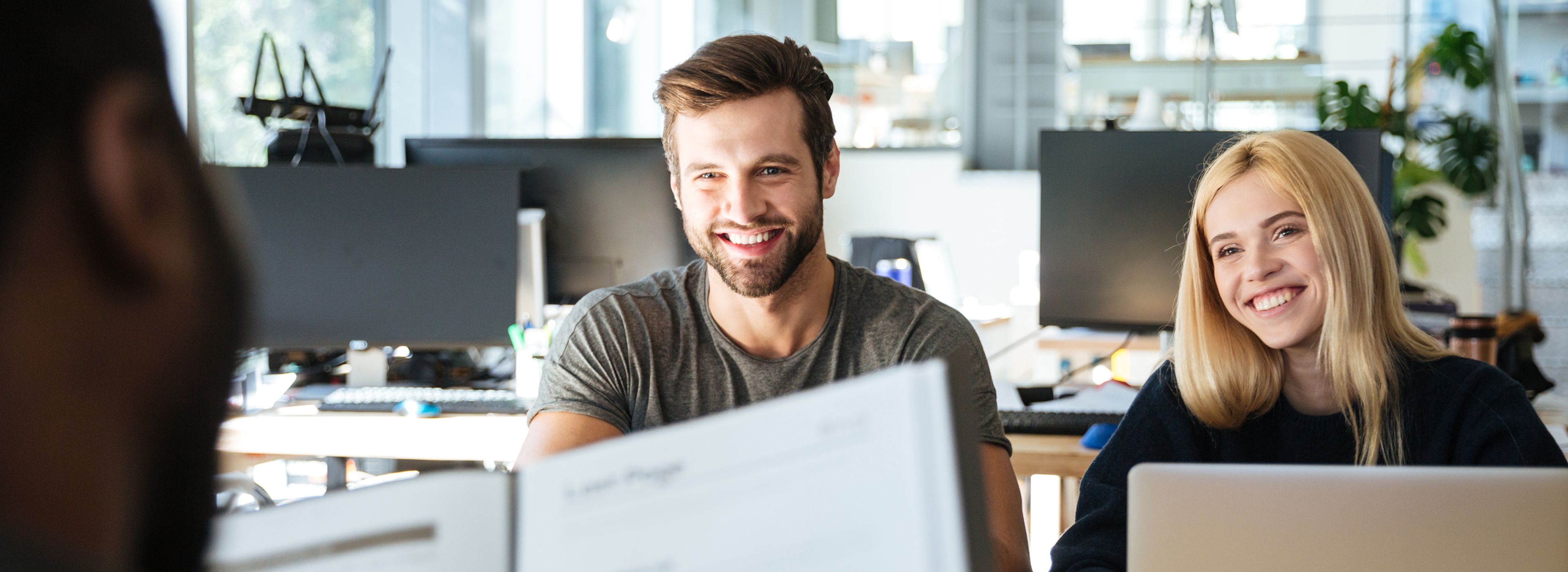 Drei Personen in einem modernen Büro, zwei von ihnen lächeln auf einen Computerbildschirm. Heller, offener Raum mit großen Fenstern und Pflanzen im Hintergrund.