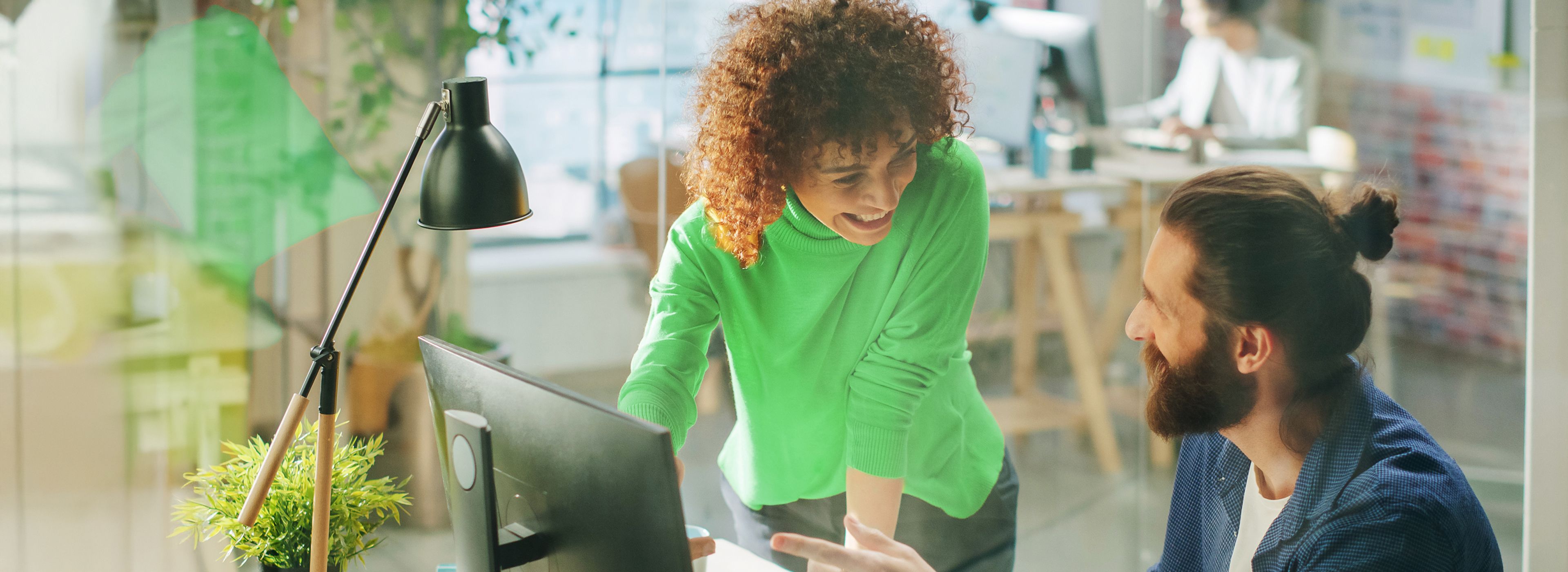 Zwei Kollegen in einem modernen Büro, eine steht und lächelt, der andere sitzt und gestikuliert – beide sind in ein Gespräch am Computer vertieft.