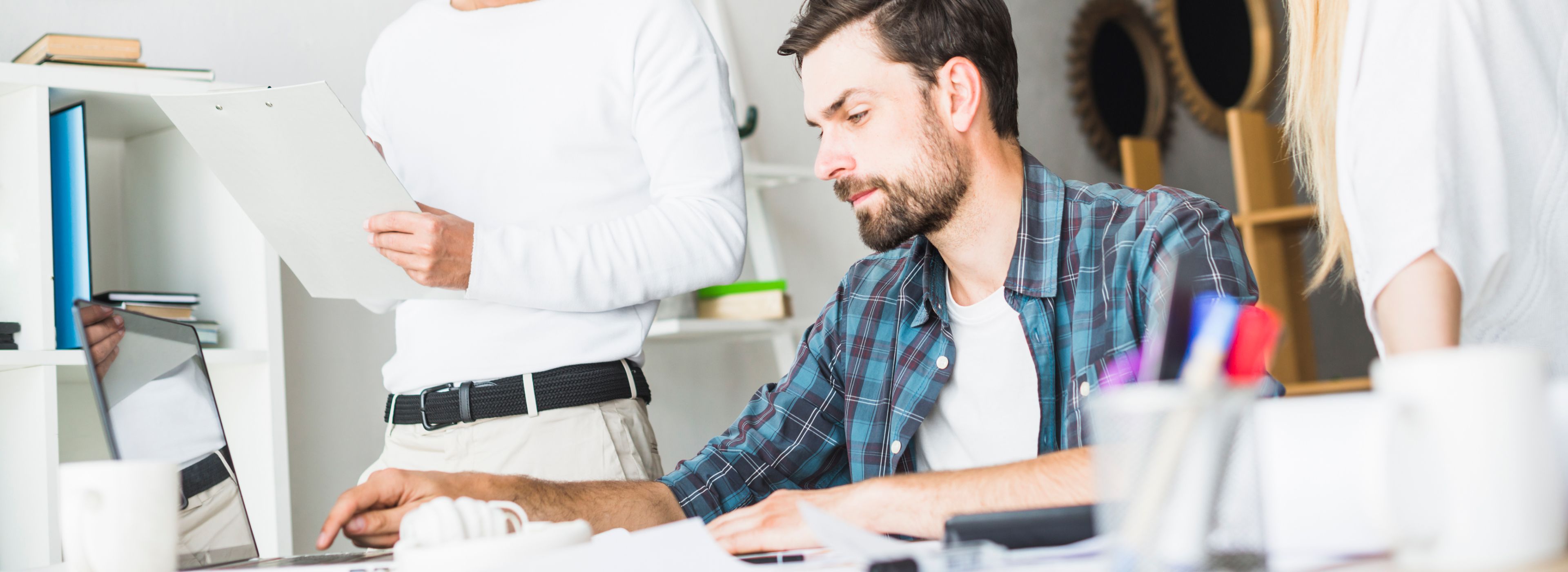 Ein Mann im karierten Hemd arbeitet an einem Computer an einem Schreibtisch, während zwei Kollegen in einem hellen Büro in der Nähe stehen und Dokumente in der Hand halten.