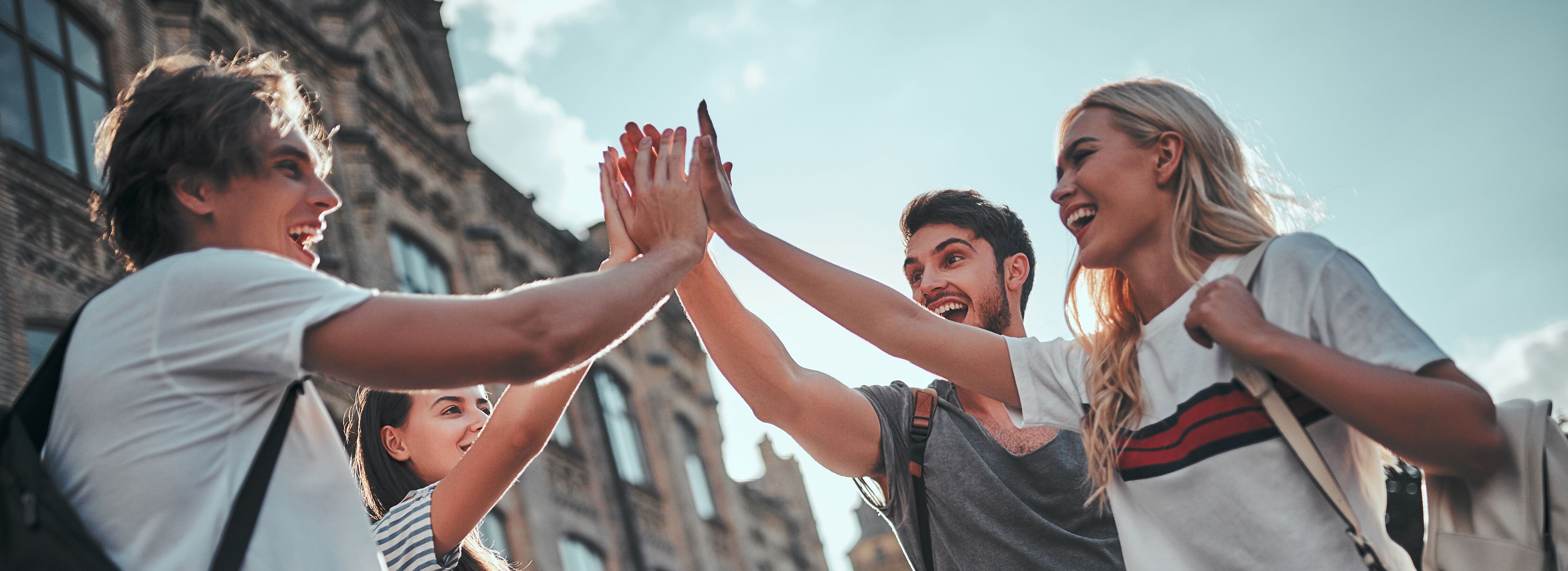 Vier Freunde lächeln und geben sich im Freien gemeinsam ein High-Five, im Hintergrund ein historisches Gebäude unter einem klaren blauen Himmel.