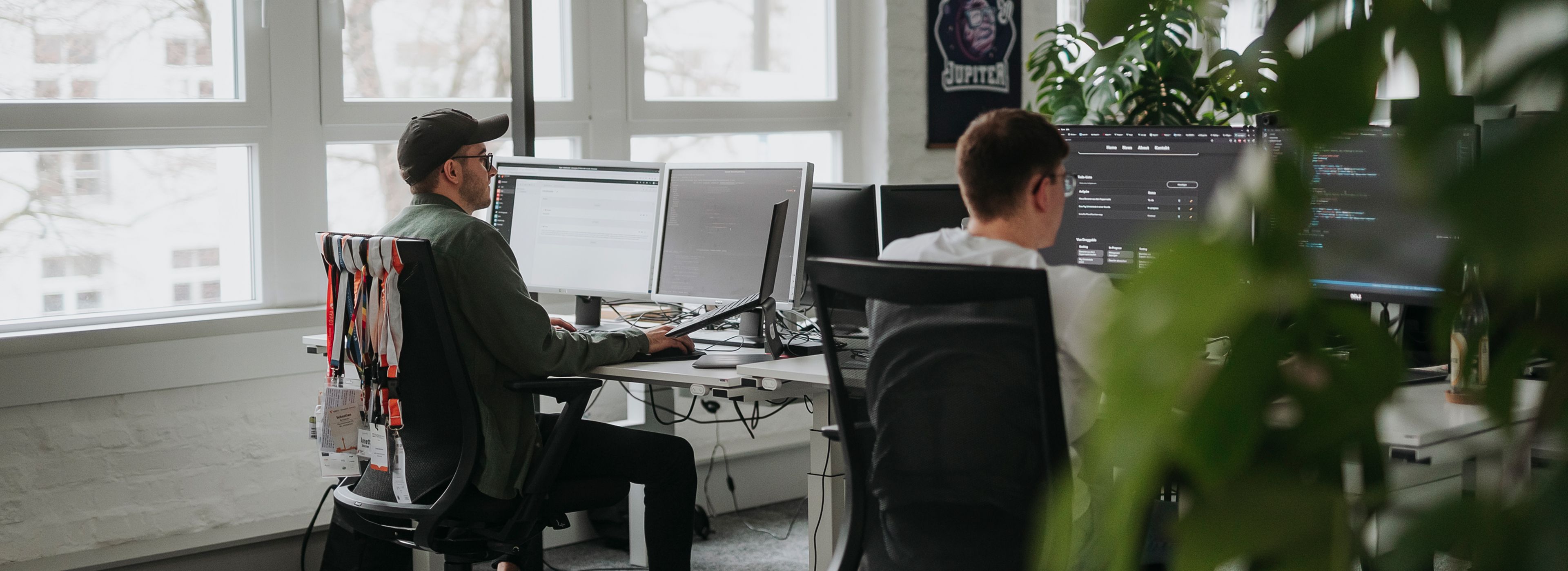 Zwei Männer arbeiten an mehreren Monitoren in einem modernen Büro mit großen Fenstern und Pflanzen im Hintergrund. Auf den Bildschirmen sind Programmier- und Interface-Anwendungen zu sehen.