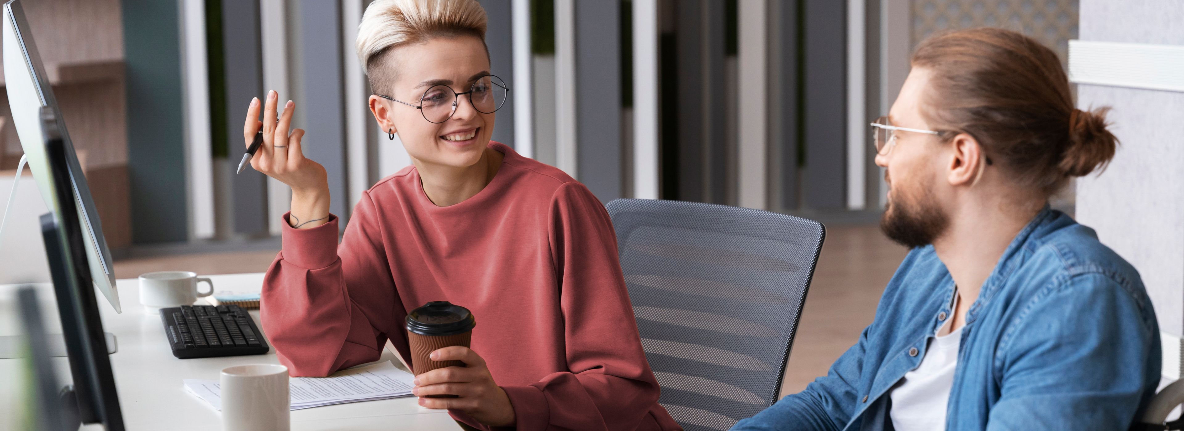 Zwei Personen sitzen an einem Schreibtisch, eine davon hält eine Kaffeetasse und einen Stift, beide sind in ein Gespräch vertieft. Auf dem Schreibtisch sind ein Computer und eine Tastatur zu sehen.