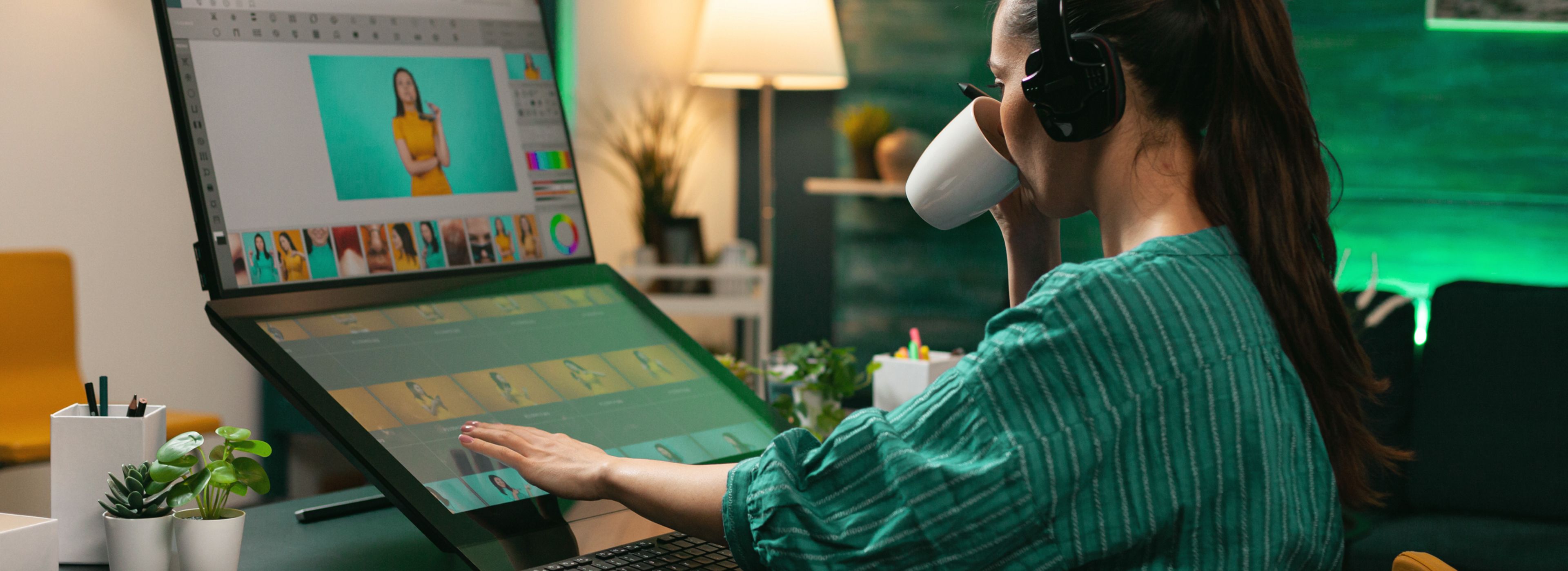 Eine Person mit Kopfhörern arbeitet an einem großen Touchscreen-Computer und bearbeitet Bilder. Dabei trinkt sie aus einer Tasse und ist von Pflanzen und Bürodekoration umgeben.