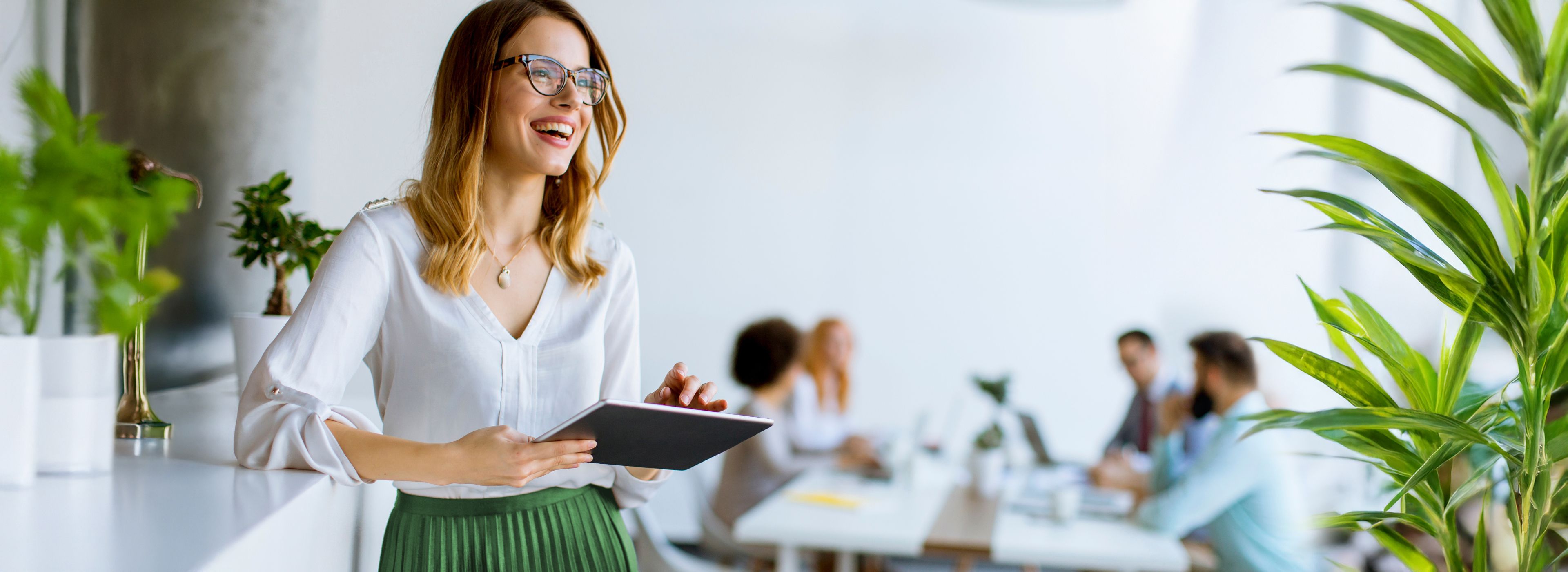 Lächelnde Frau mit Brille hält ein Tablet in der Hand und steht nahe einem Schreibtisch mit Pflanzen; im Hintergrund arbeitet eine Gruppe von Personen.