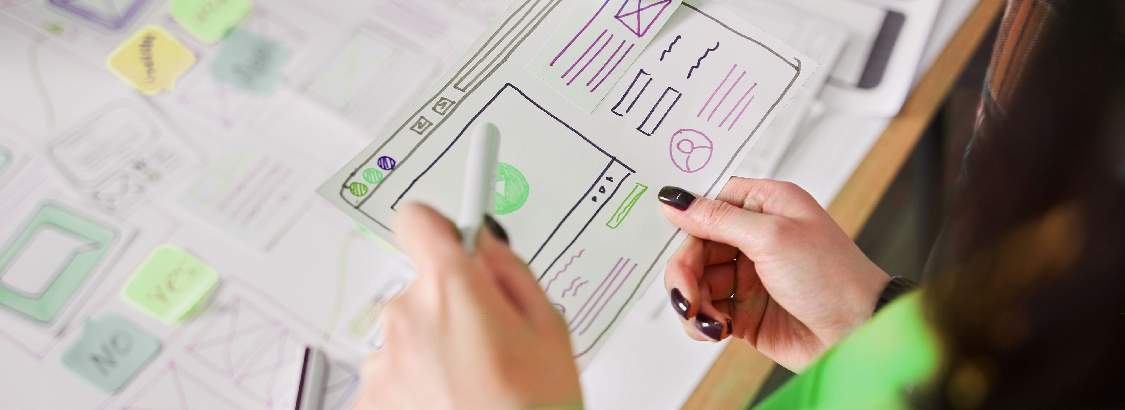 Eine Person hält ein Blatt mit Skizzen von Website-Wireframes, umgeben von Haftnotizen und Designmaterialien auf einem Tisch.