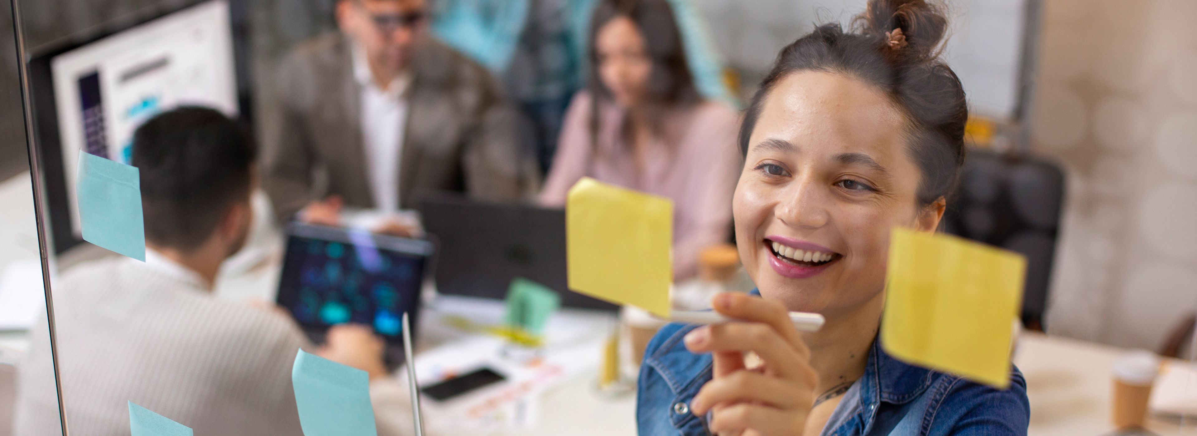 Lächelnde Frau in einer Jeansjacke klebt eine Notiz auf eine Glaswand in einem Büro, während Kollegen im Hintergrund an Laptops arbeiten.