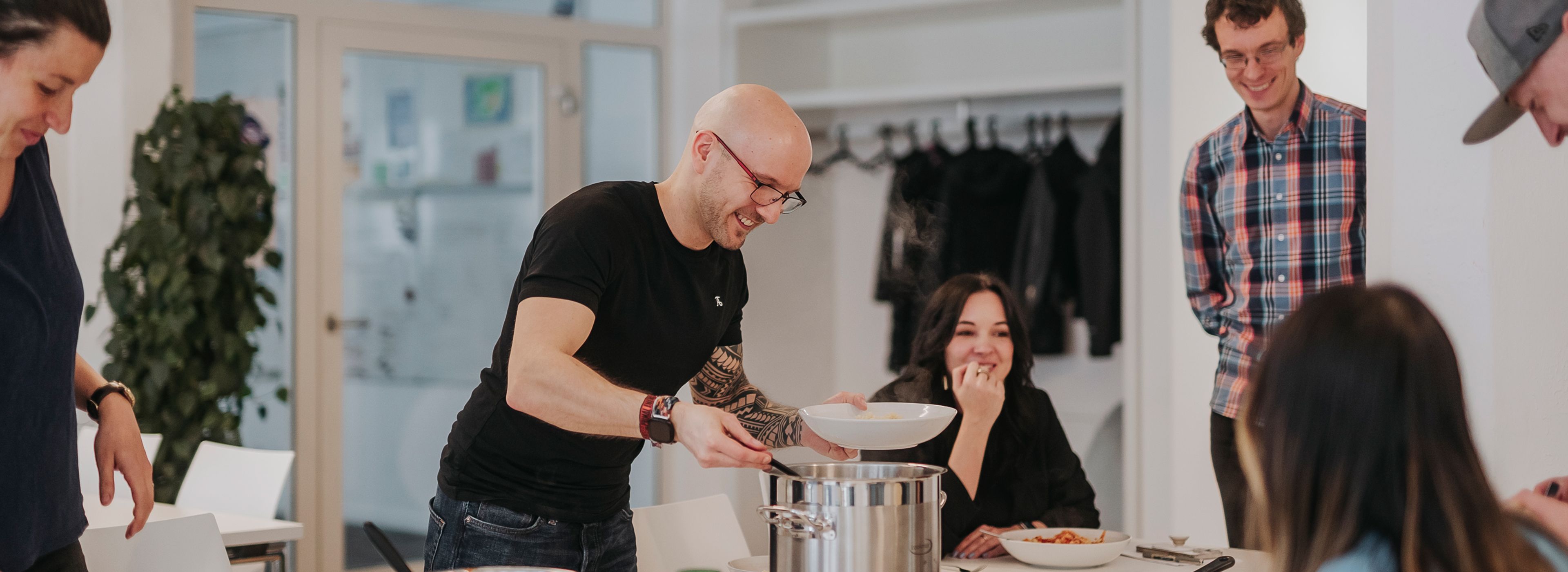 Eine Gruppe von Personen befindet sich in einer Küche, während ein Mann Essen aus einem großen Topf in eine Schüssel serviert. Sie lächeln und unterhalten sich miteinander.