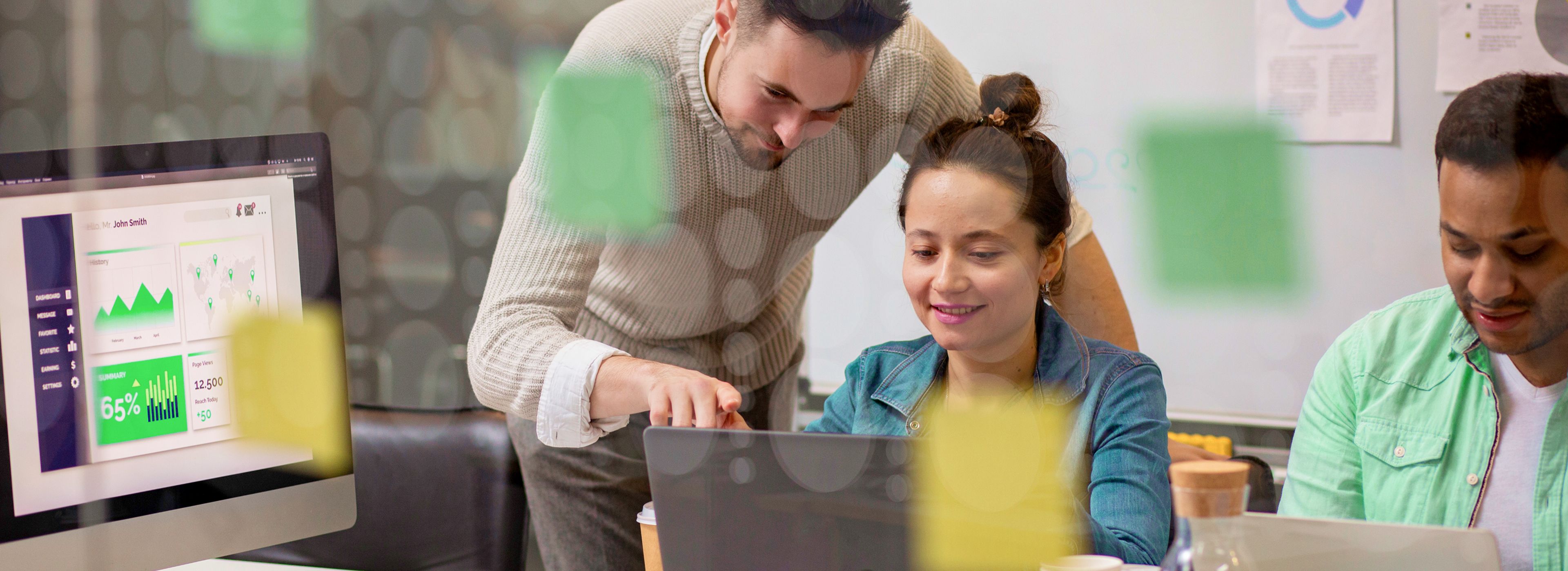 Drei Personen arbeiten gemeinsam in einem Büro, wobei eine Person auf den Laptopbildschirm zeigt. Im Hintergrund zeigt ein Computer Diagramme an.