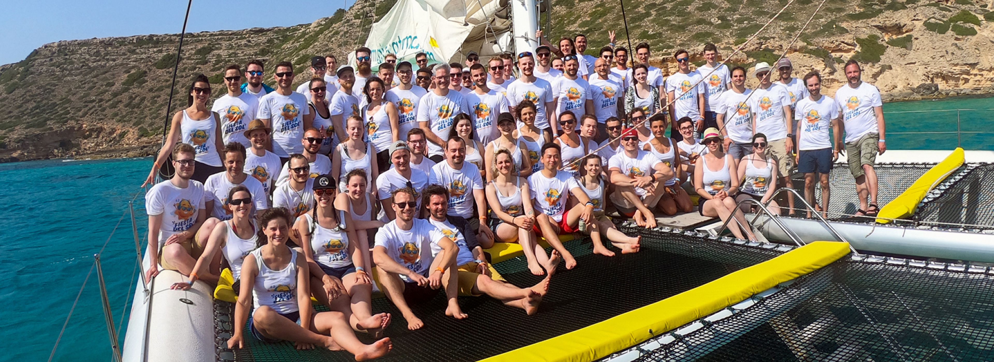 Eine große Gruppe von Personen in einheitlichen Shirts posiert auf einem Katamaran, im Hintergrund felsige Klippen und blaues Wasser.