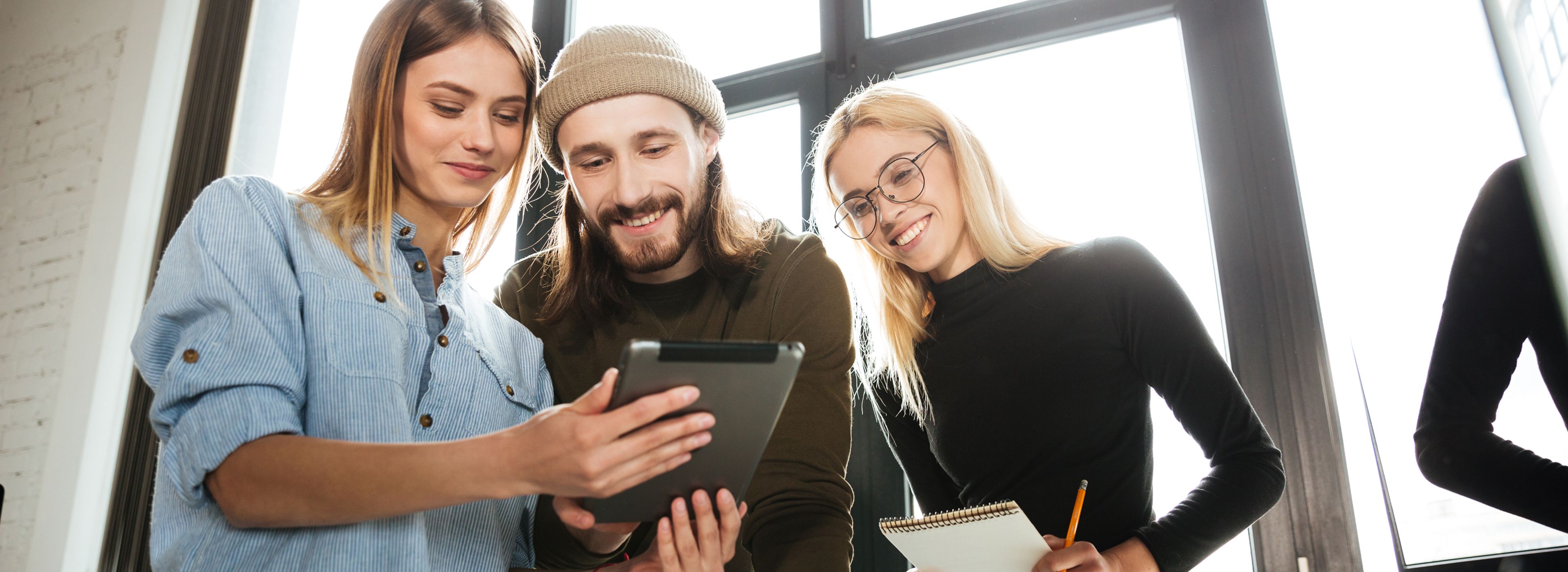 Drei junge Kollegen (Frau, Mann, Frau) in einem Büro schauen gemeinsam auf ein Tablet, während die Frau rechts einen Notizblock und einen Stift in der Hand hält.