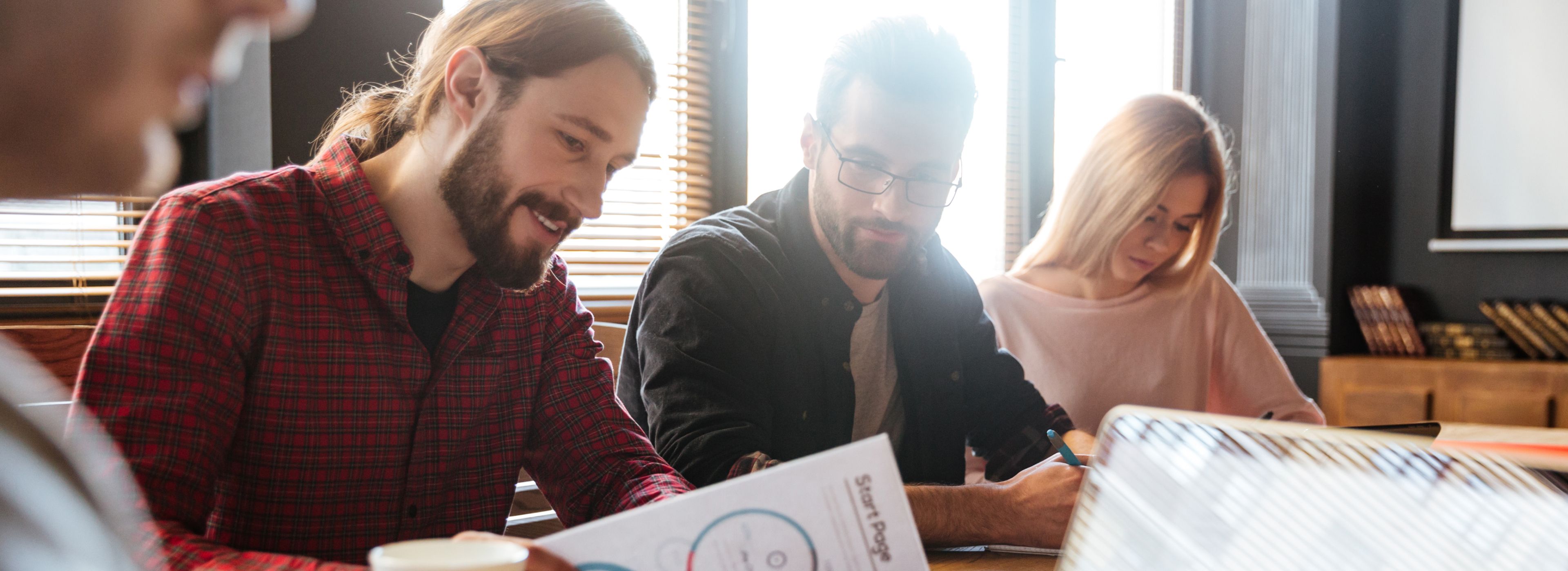 Drei Kollegen (Mann, Mann, Frau) sitzen zusammen an einem Tisch in einem Büro. Der Mann ganz links im karierten Hemd lächelt, während er ein Dokument betrachtet, ein anderer schreibt Notizen, und eine Frau im Hintergrund arbeitet konzentriert.