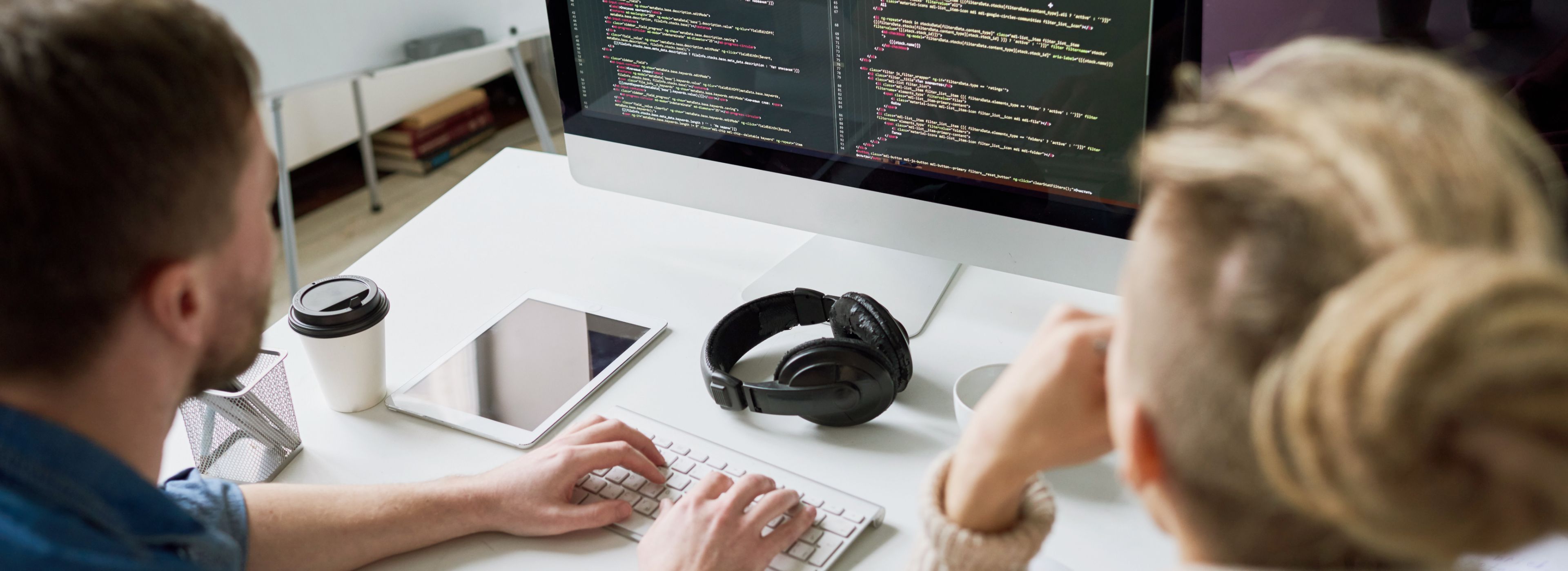 Zwei Personen arbeiten an einem Schreibtisch mit einem Computer, auf dem Code angezeigt wird. In der Nähe befinden sich ein Tablet, Kopfhörer und eine Kaffeetasse.
