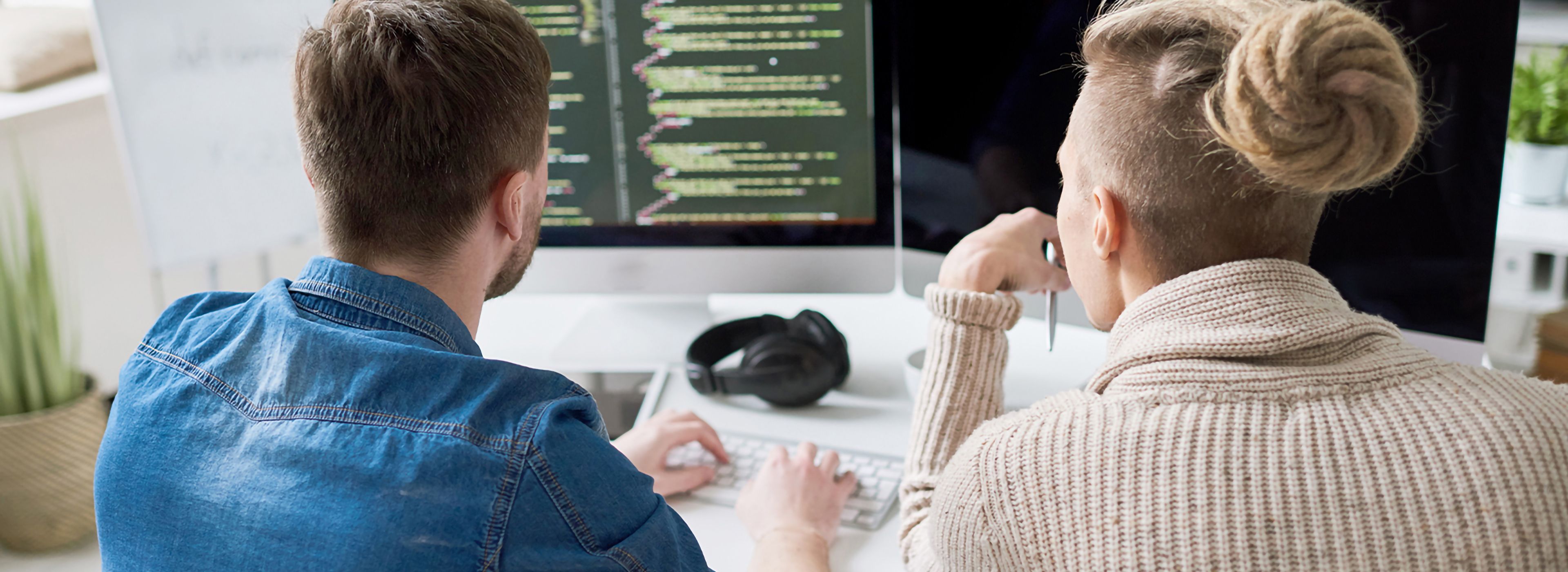 Zwei Personen sitzen an einem Schreibtisch und arbeiten an Code, der auf einem Computermonitor angezeigt wird. Eine trägt ein Jeanshemd, die andere einen beigefarbenen Pullover und hat die Haare zu einem Dutt gebunden.