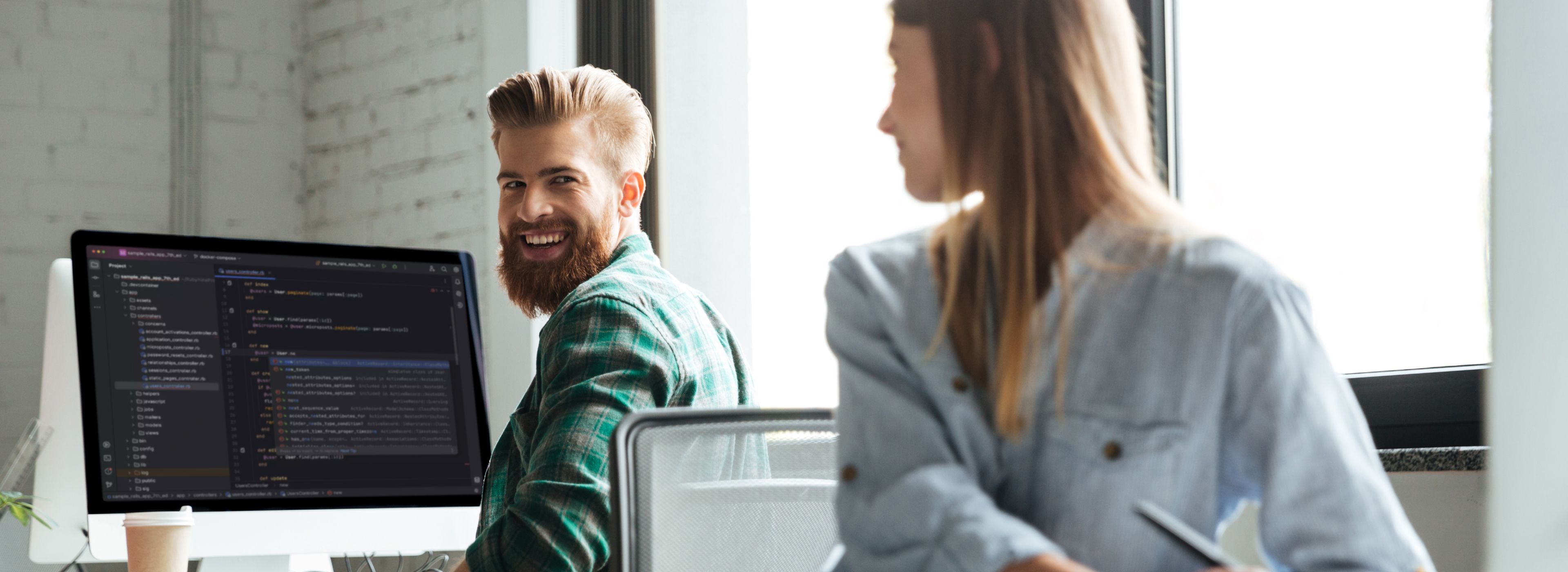 Ein Mann mit Bart und grünem Flanellhemd sitzt an einem Schreibtisch und schaut lächelnd über die Schulter zu einer Kollegin. Auf seinem Bildschirm ist Code in einer Entwicklungsumgebung zu sehen. Im Hintergrund ist ein helles Fenster erkennbar.