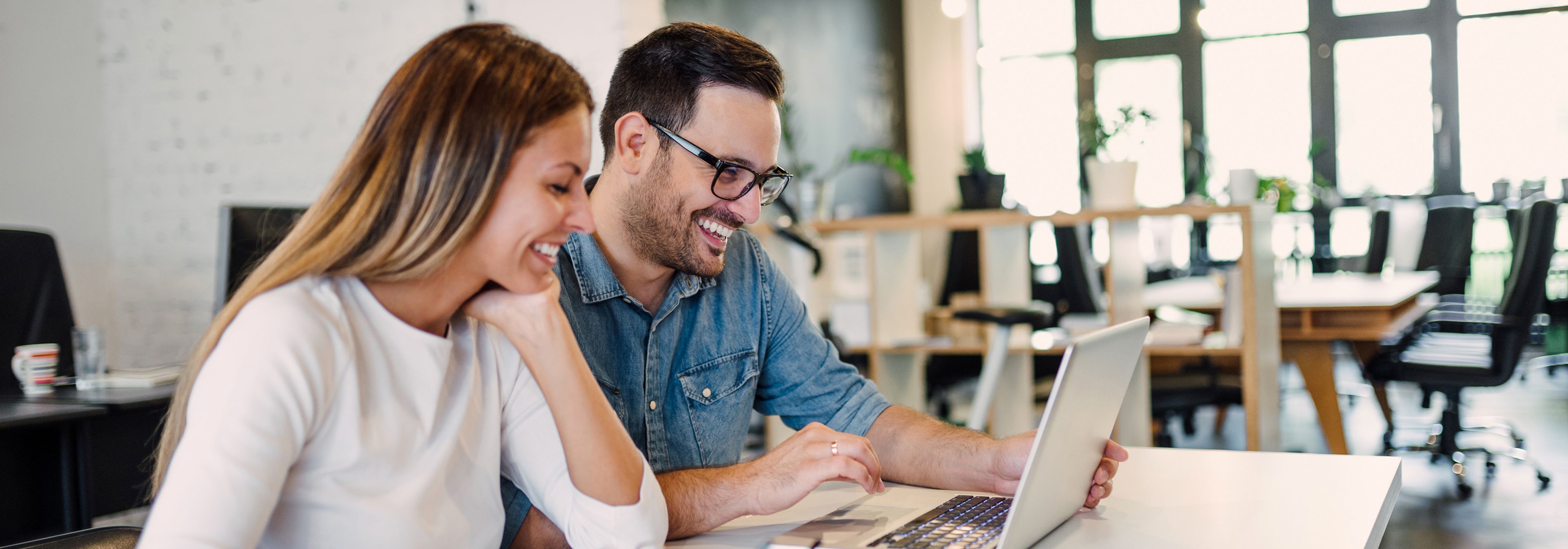 Ein Mann und eine Frau lächeln und schauen auf einen Laptop in einem modernen Büro mit Schreibtischen und großen Fenstern im Hintergrund.