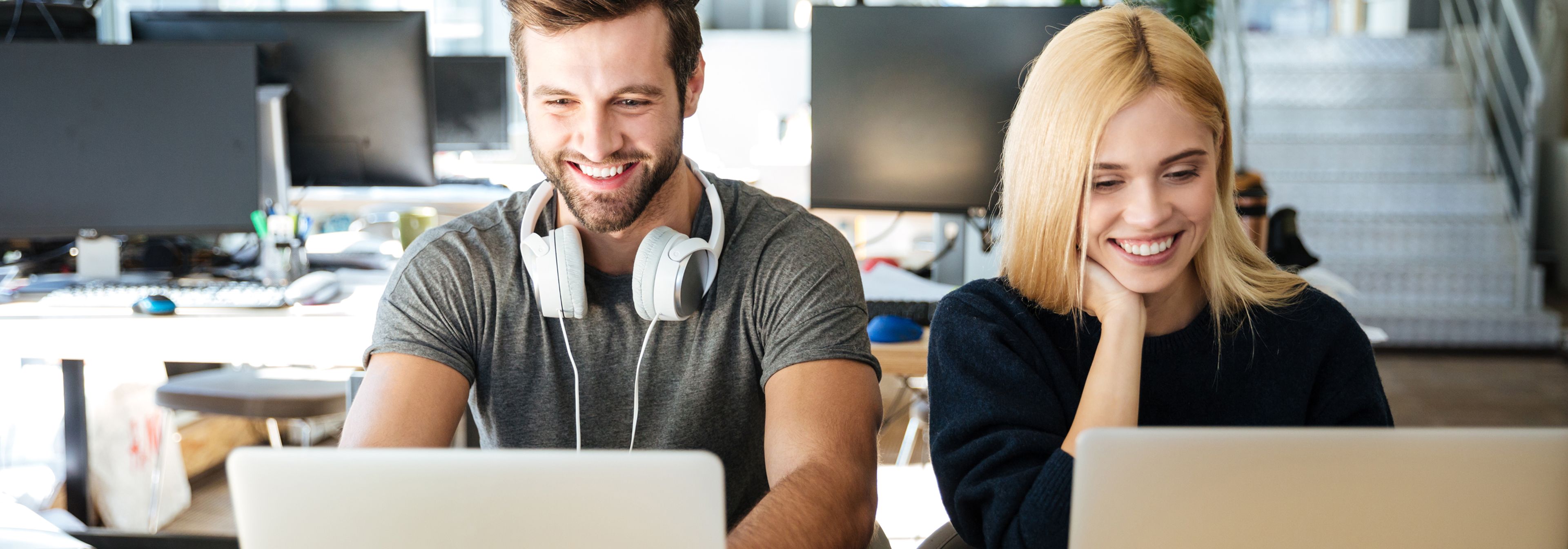 Zwei Personen sitzen nebeneinander und lächeln, während sie an Laptops in einem hellen, modernen Büro arbeiten. Eine von ihnen trägt Kopfhörer um den Hals.