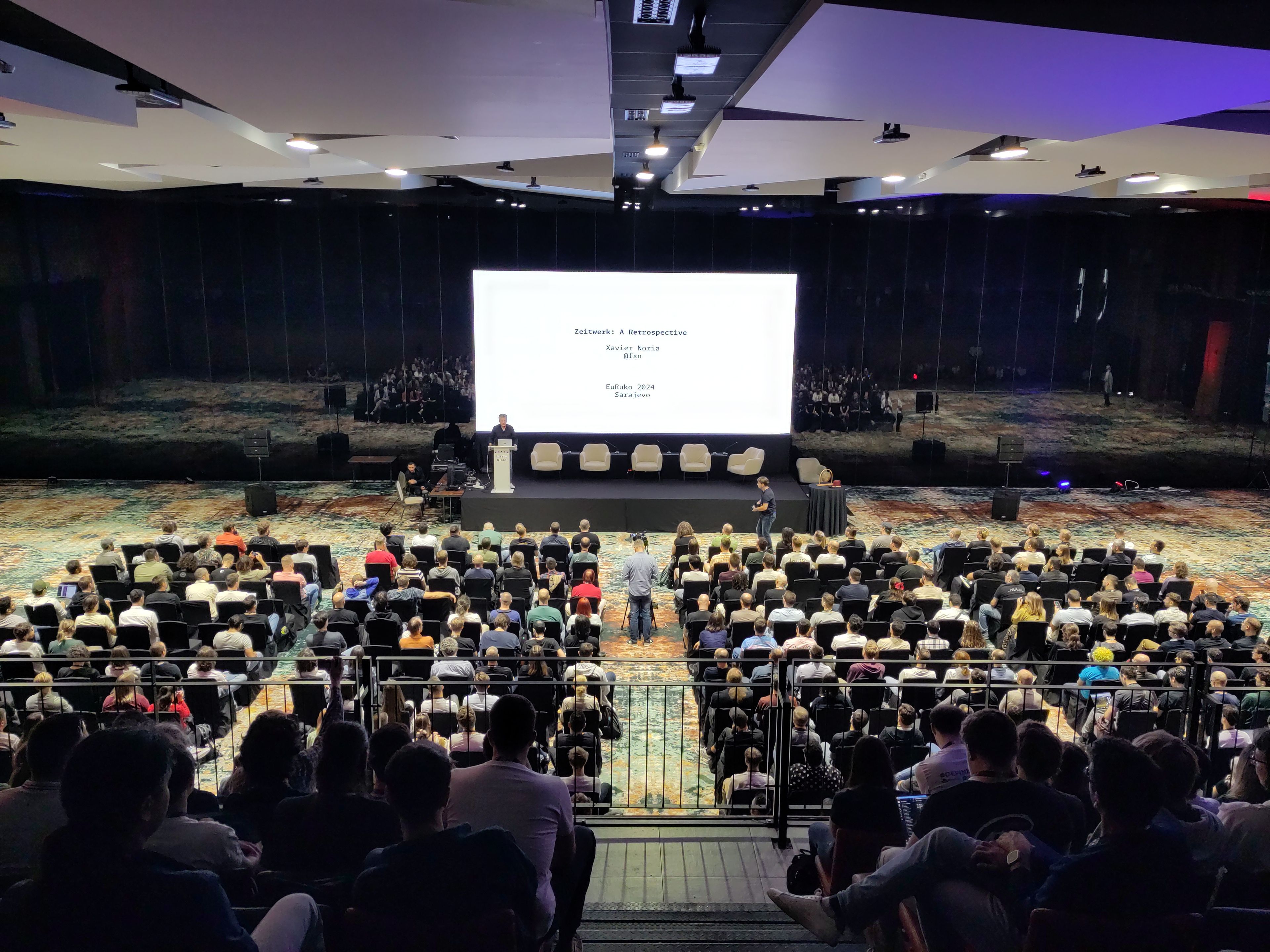 Eine Konferenzveranstaltung mit zahlreichen Teilnehmern, die in einem großen Saal sitzen. Auf einer Bühne spricht eine Person vor einer großen Leinwand, die den Vortragstitel „Zeitwerk: A Retrospective“ zeigt. Die Umgebung ist modern und professionell mit reflektierenden Wänden und hochwertiger Beleuchtung.