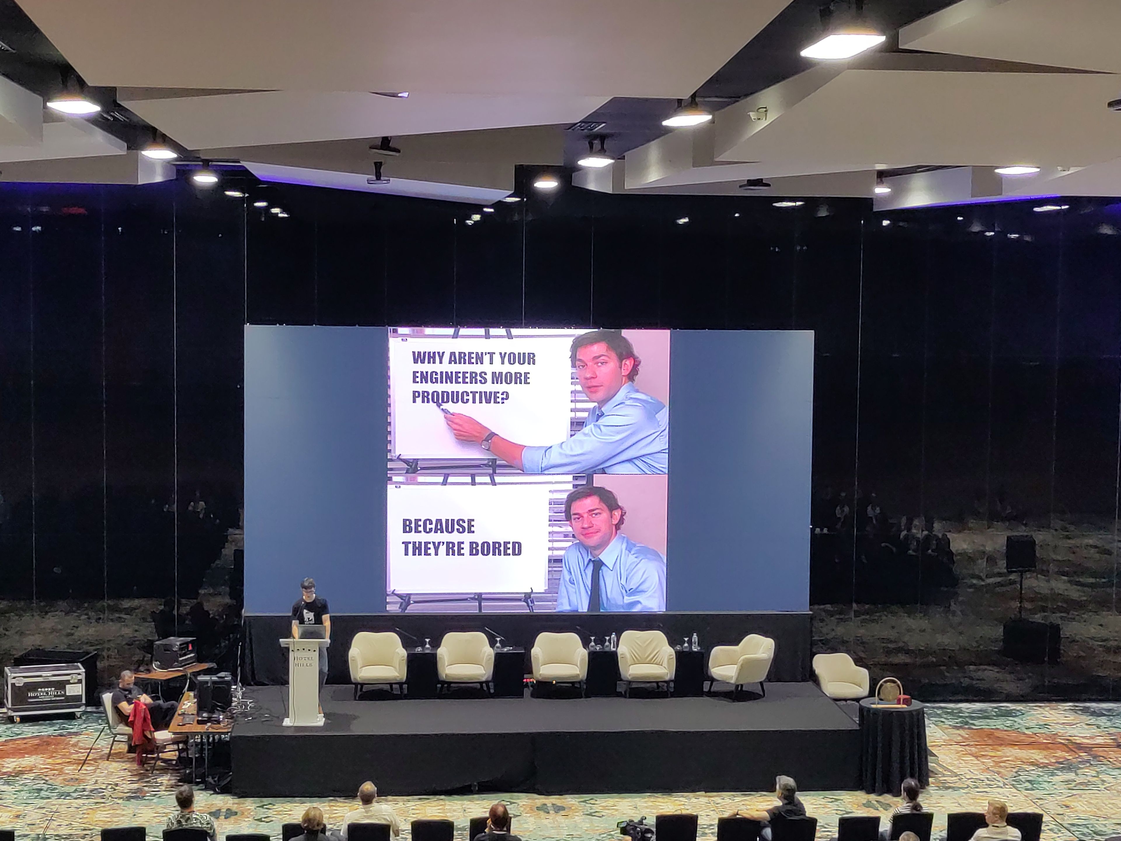 Ein Vortragssaal mit einer großen Leinwand, auf der ein humorvolles Meme über Produktivität in der Softwareentwicklung gezeigt wird. Eine Person steht am Rednerpult und spricht vor einem Publikum, das auf Reihen von Stühlen sitzt. Der Raum ist modern gestaltet und mit professionellem Equipment ausgestattet.