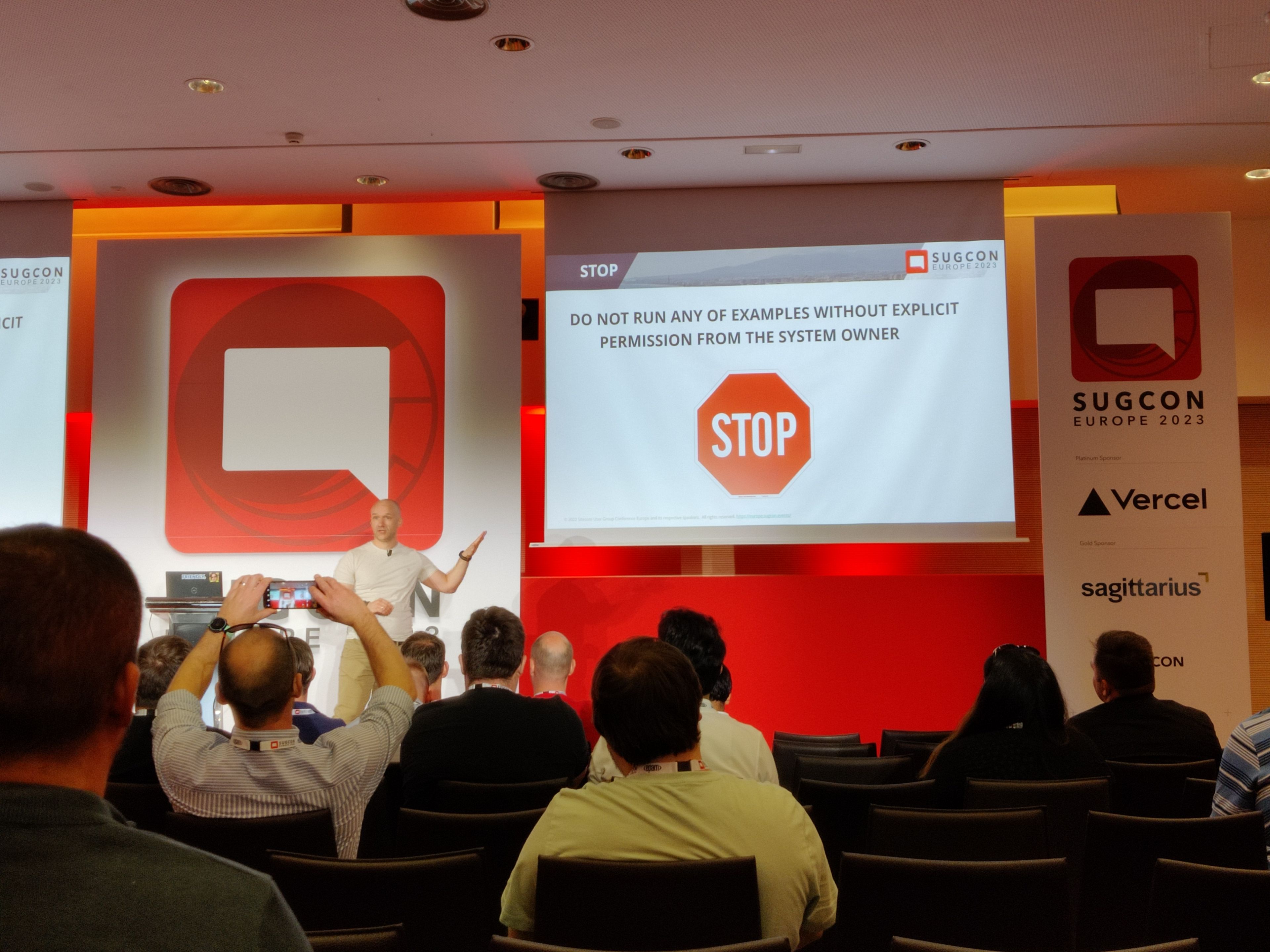 Ein Vortrag auf der SUGCON Europe 2023. Ein Redner präsentiert vor einer Leinwand mit der Aufschrift „Do not run any of examples without explicit permission from the system owner“. Das Publikum hört zu, während einige Teilnehmer Fotos machen.