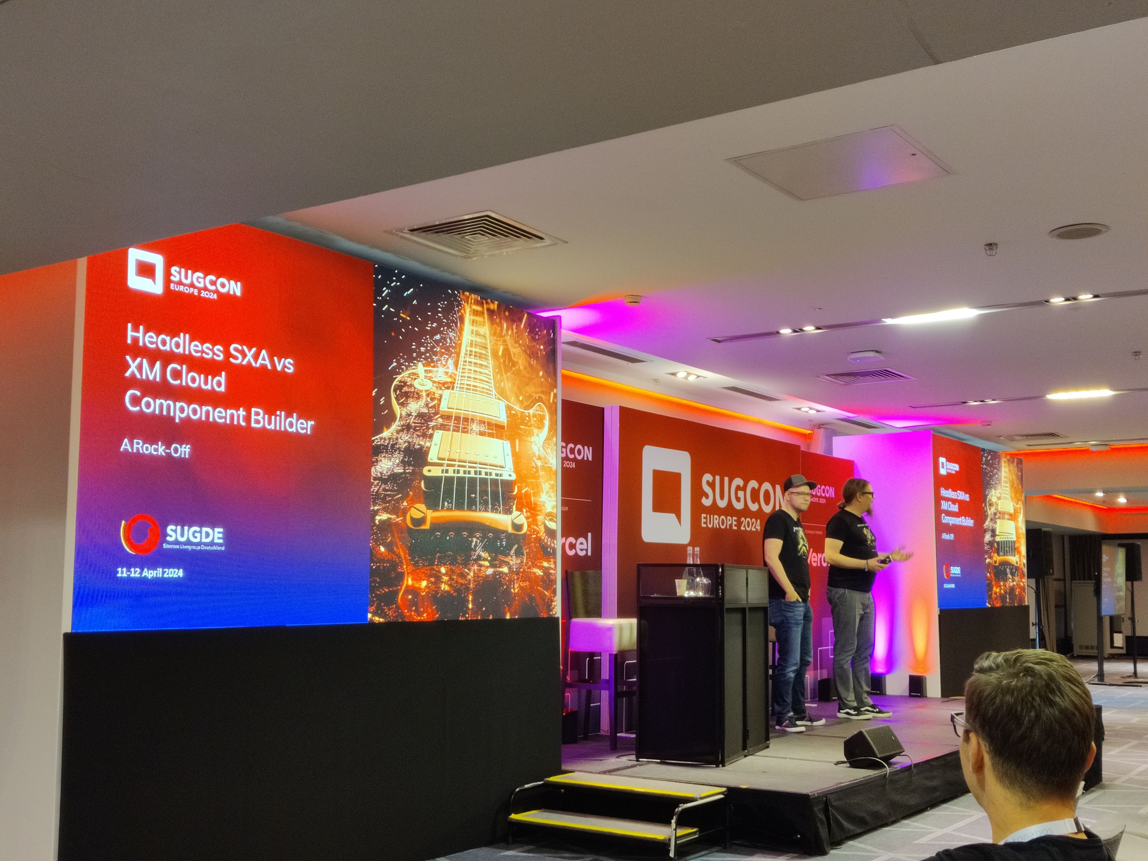 Eine Präsentation auf der SUGCON Europe 2024. Links ist eine große Leinwand mit einer Ankündigung zu sehen: „Headless SXA vs XM Cloud Component Builder – A Rock-Off“, begleitet von einem Bild einer E-Gitarre in feurigen Effekten. Rechts auf der Bühne stehen zwei Sprecher, die in lockerer Atmosphäre das Thema präsentieren. Im Hintergrund und auf der Bühne sind das Logo und Branding der SUGCON Europe 2024 gut sichtbar. Im Vordergrund sitzt ein Teilnehmer, der der Präsentation aufmerksam folgt. Die Bühne ist in einem modernen Konferenzraum mit farbiger Beleuchtung aufgebaut.