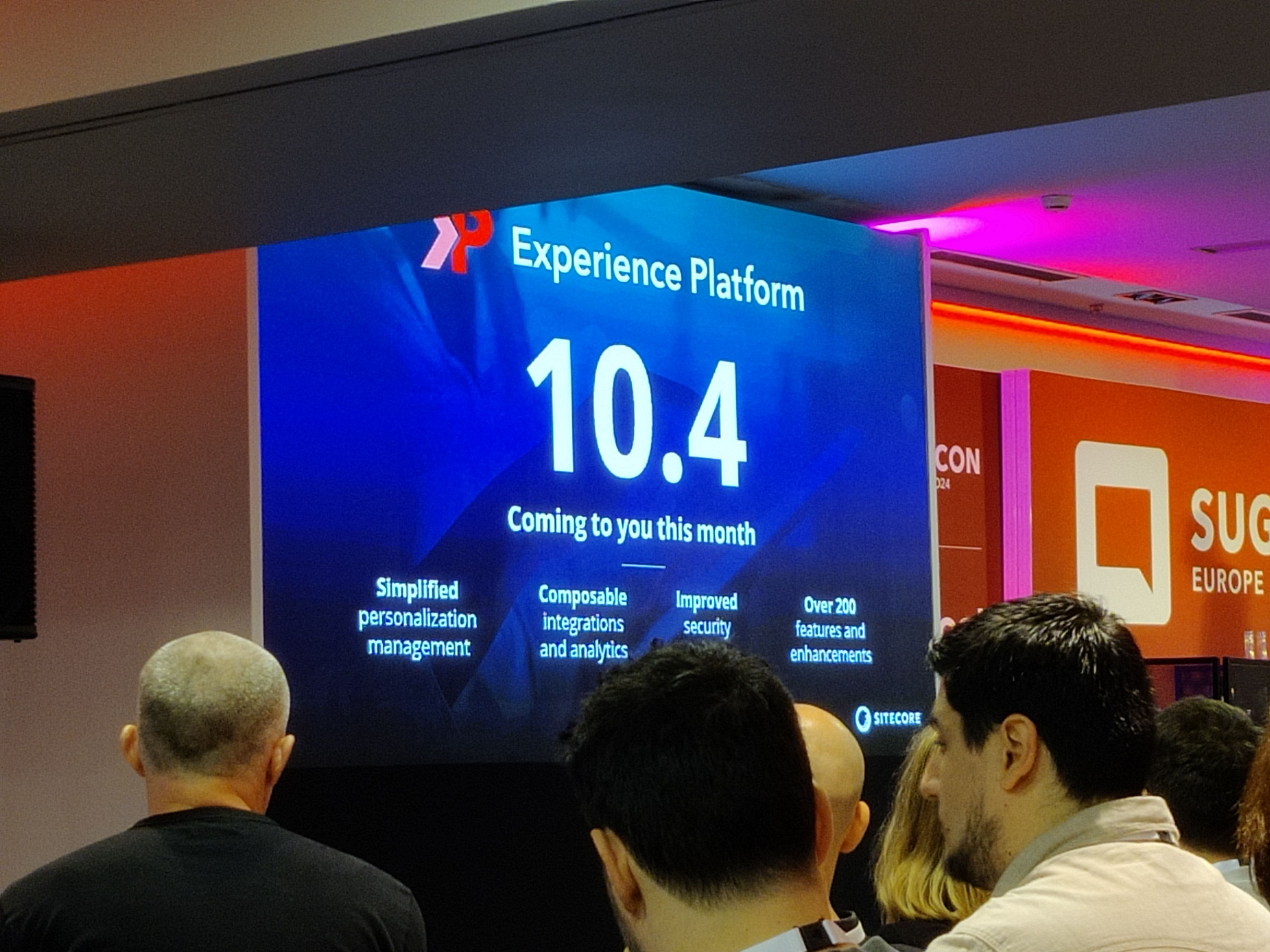 Eine Präsentation auf einer Konferenz. Im Vordergrund sind Teilnehmer zu sehen, die der Präsentation zuhören. Auf der Leinwand wird die Version „10.4“ der „Experience Platform“ angekündigt, die in diesem Monat verfügbar sein soll. Hervorgehoben werden Funktionen wie „vereinfachtes Personalisierungsmanagement“, „komponierbare Integrationen und Analysen“, „verbesserte Sicherheit“ sowie „über 200 neue Funktionen und Verbesserungen“. Rechts im Bild sind das Logo und Branding der SUGCON Europe 2024 erkennbar.