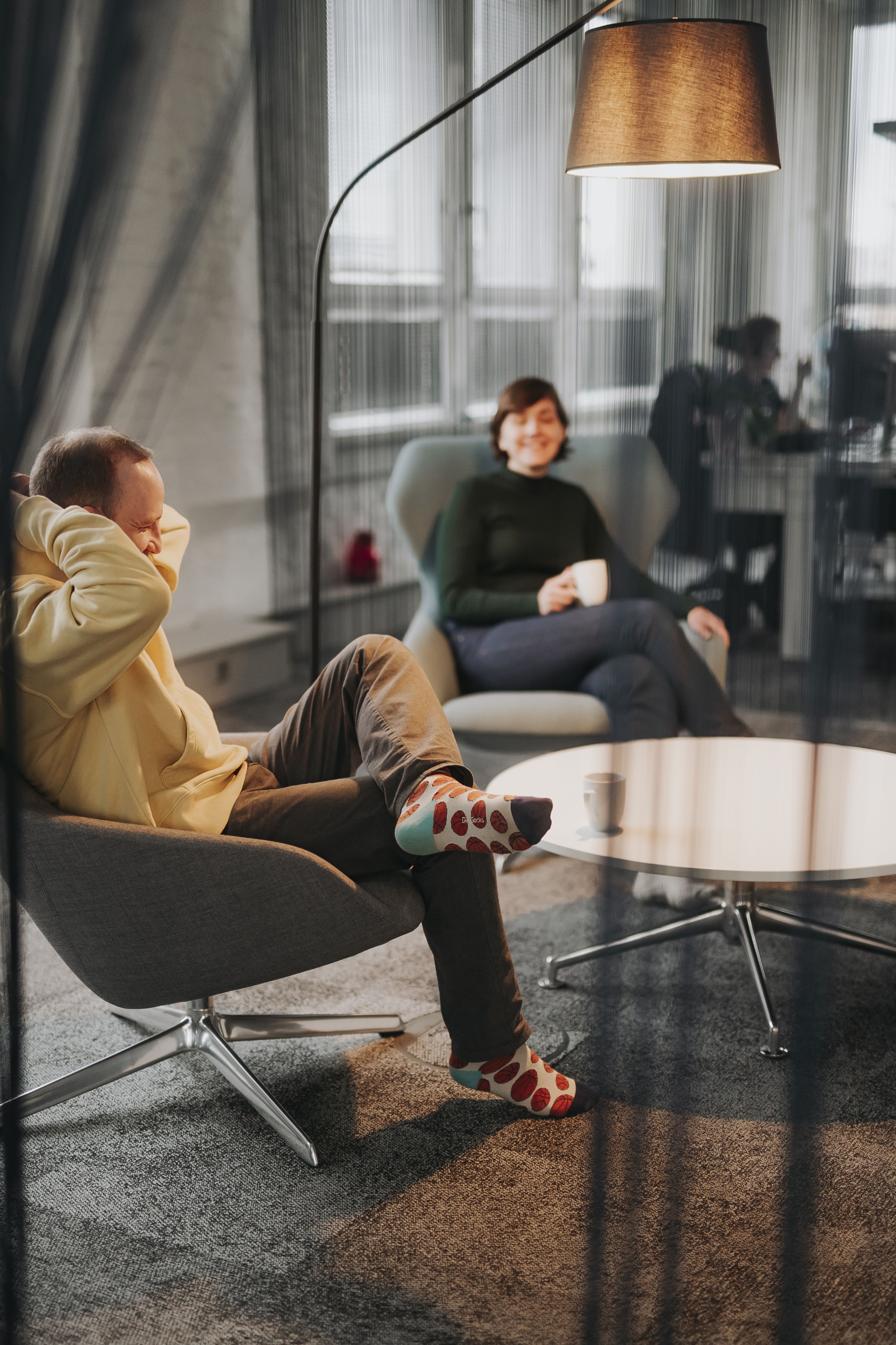 Zwei Personen entspannen sich in modernen Sesseln und unterhalten sich unter einer Stehlampe. Eine hält eine Tasse in der Hand, die andere trägt bunte Socken. Im Hintergrund ist ein Büro zu sehen.