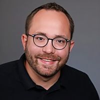 Ein lächelnder Mann mit Brille und Bart trägt ein schwarzes Poloshirt und steht vor einem schlichten grauen Hintergrund.