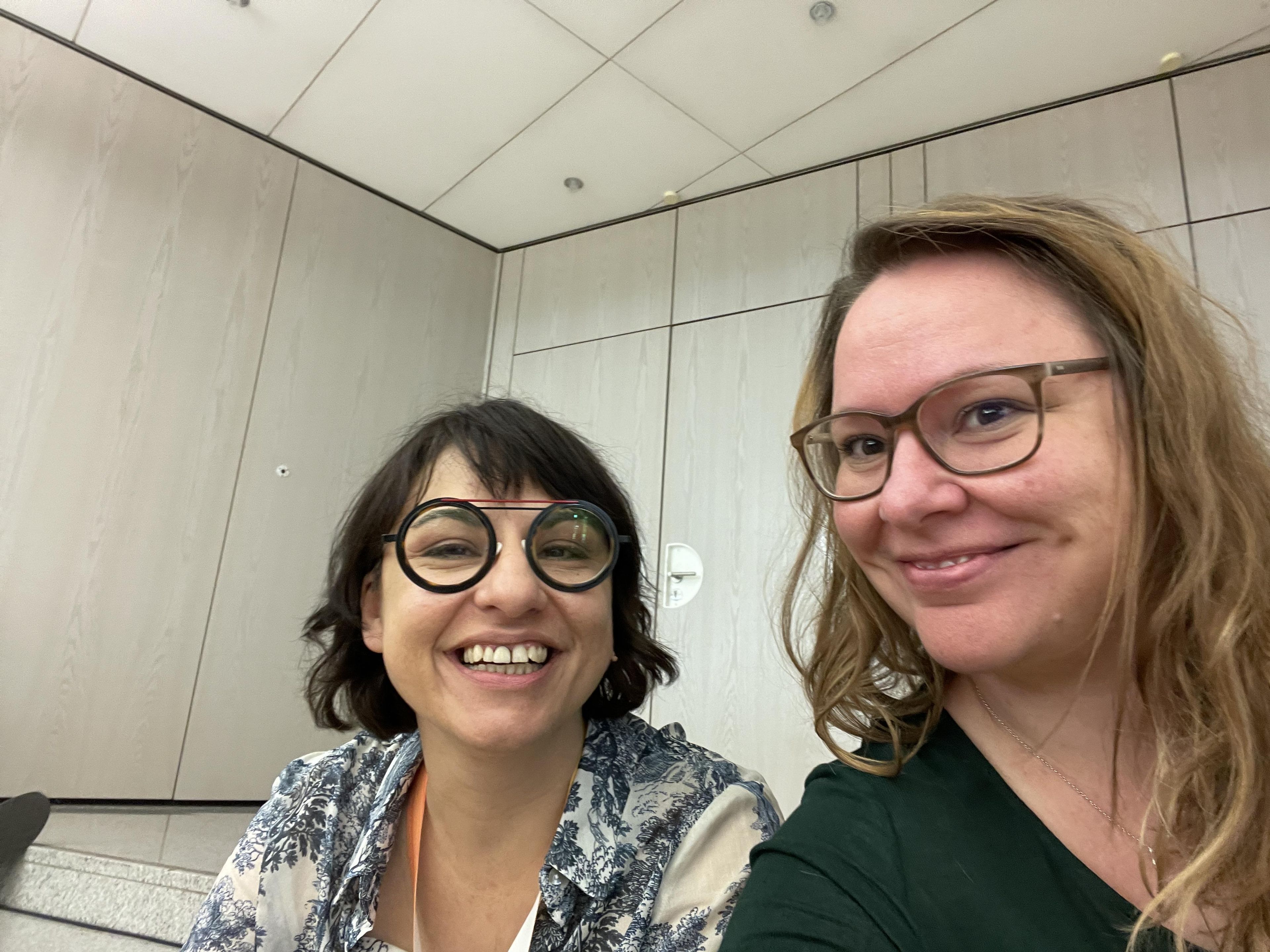 Zwei Frauen lächeln in einem Raum mit hellem Holzpaneel. Beide tragen legere Kleidung und Brillen.