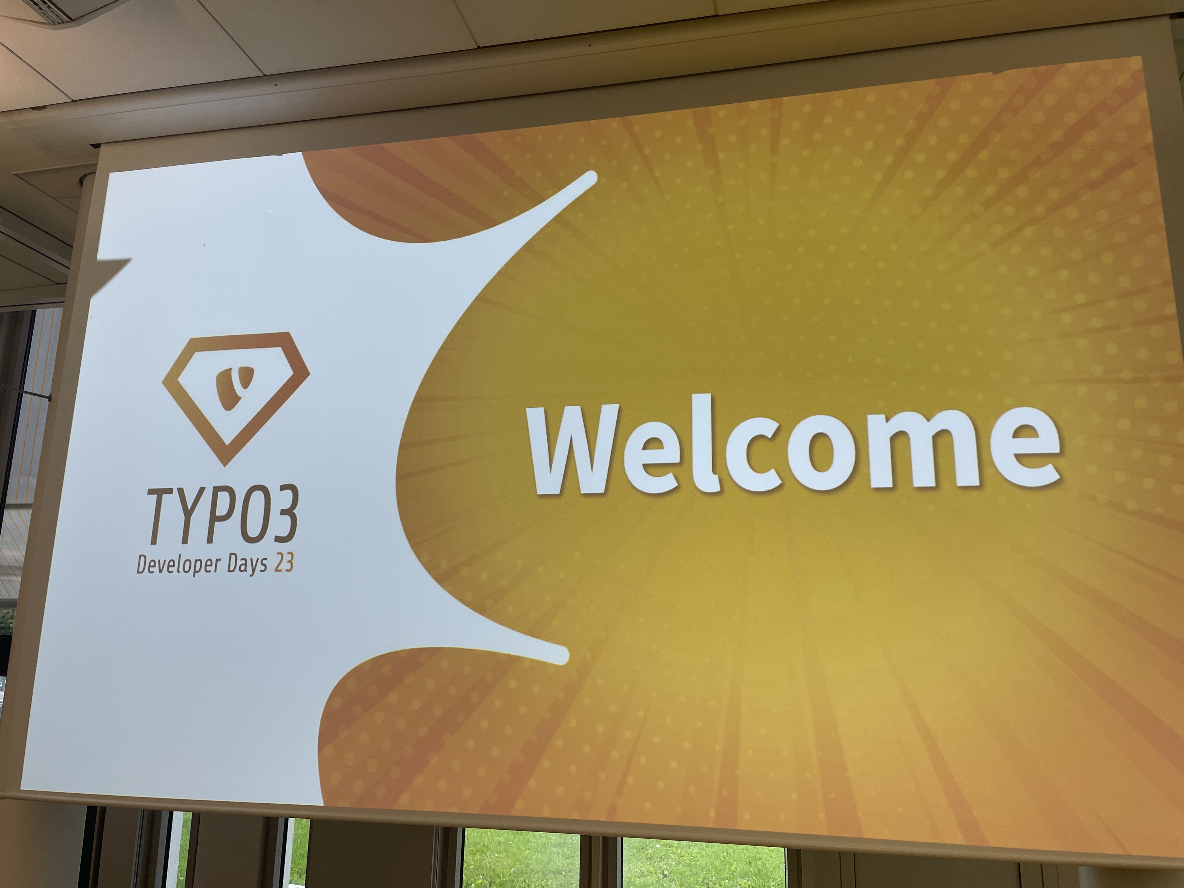Leinwand mit dem Logo „TYPO3 Developer Days 23“ und dem Text „Welcome“ auf einem orangefarbenen Hintergrund mit weißem Strahlenmotiv.