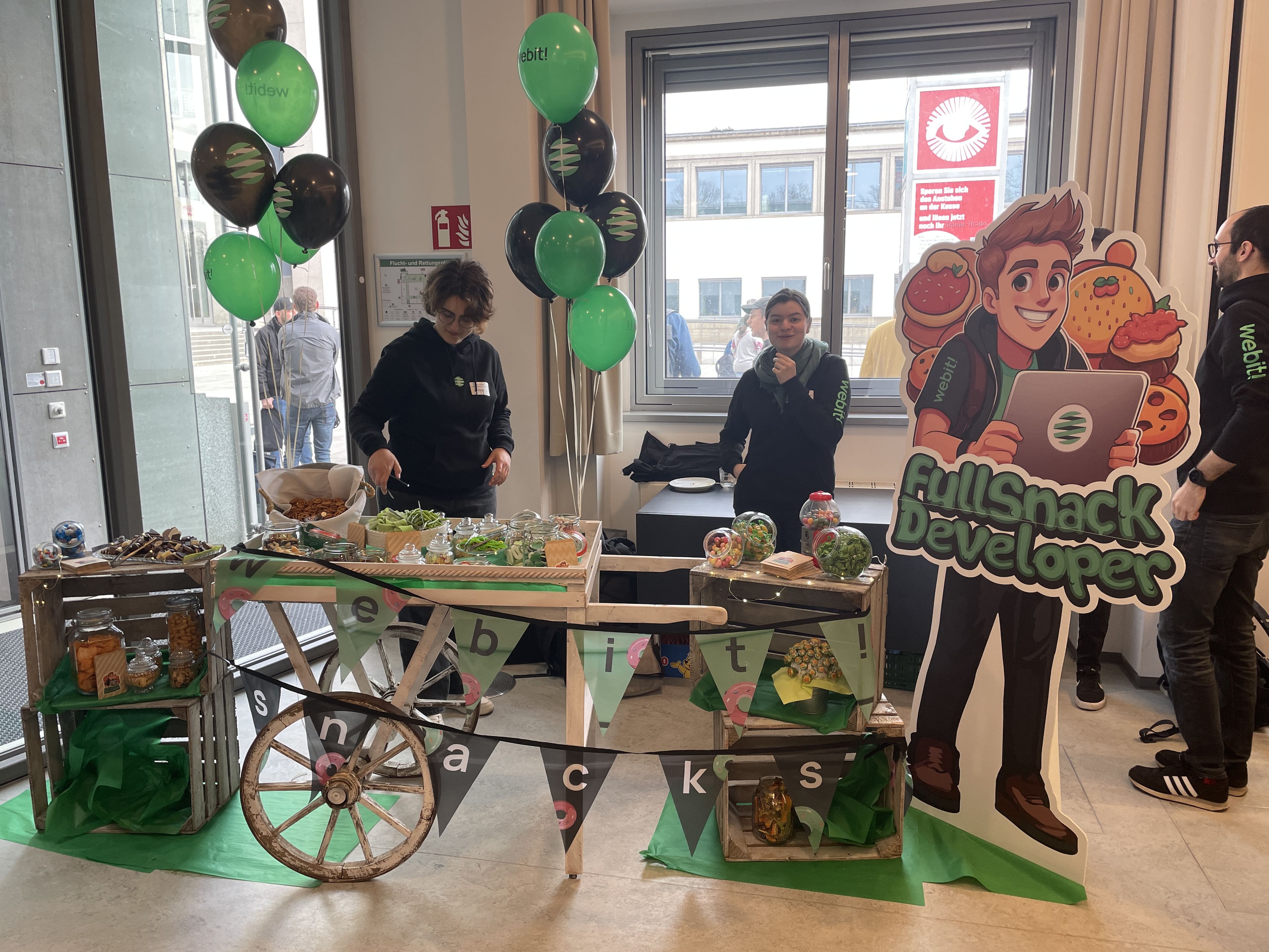 Eine Snack-Stand mit grüner und schwarzer Dekoration, Luftballons und Bannern mit der Aufschrift „webit!“. Zwei Personen stehen hinter dem Stand, der mit Gläsern voller Snacks, Schalen mit Knabbereien und Dekorationsartikeln ausgestattet ist. Daneben befindet sich eine große, humorvolle Aufstellfigur mit der Aufschrift „FullSnack Developer“, die einen Entwickler mit Laptop und Essenssymbolen darstellt. Im Hintergrund ist ein Fenster mit Blick nach draußen zu sehen.