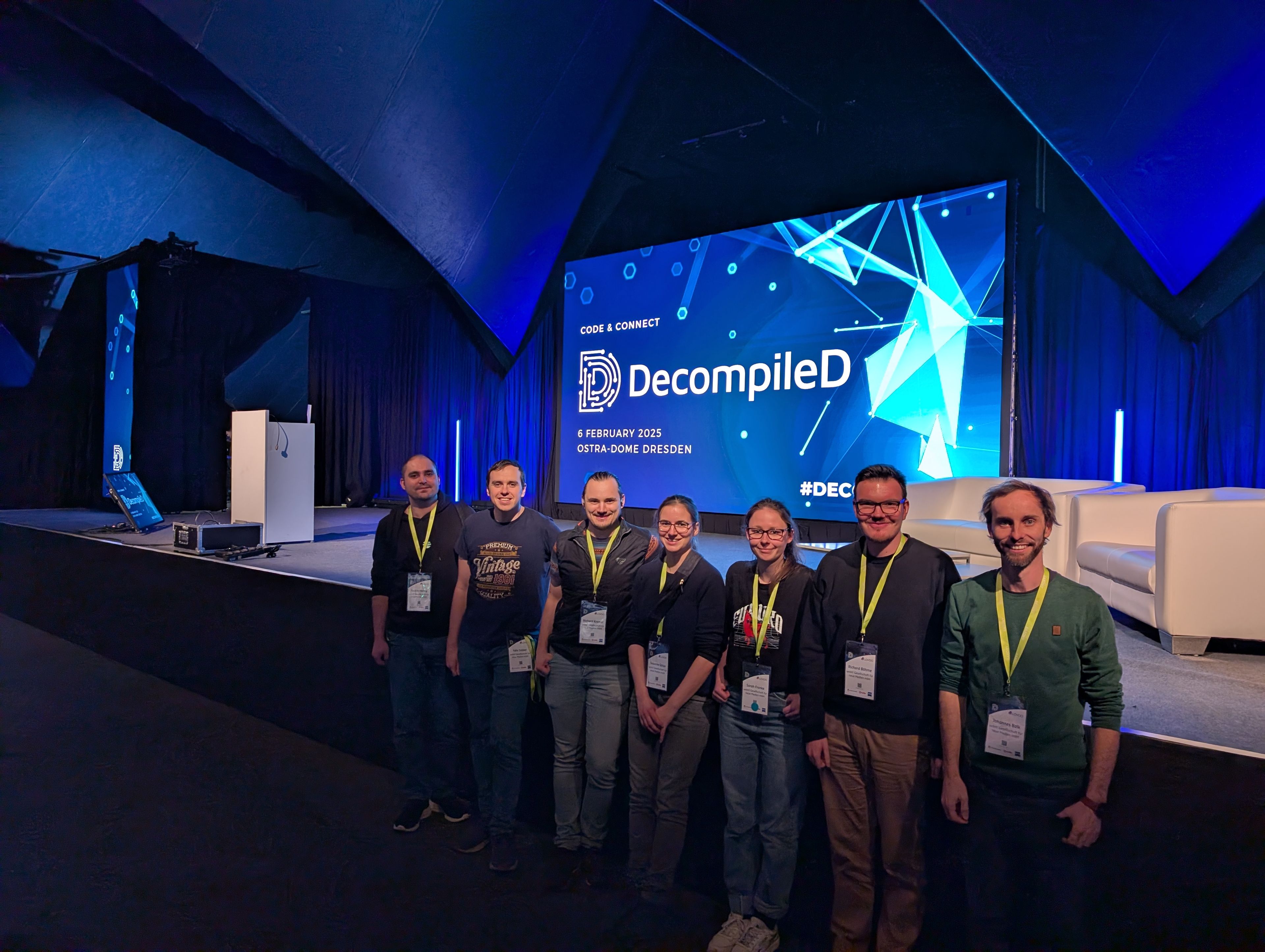 Eine Gruppe von Personen mit Lanyards steht vor einer Bühne mit einem „DecompileD“-Konferenz-Hintergrund.